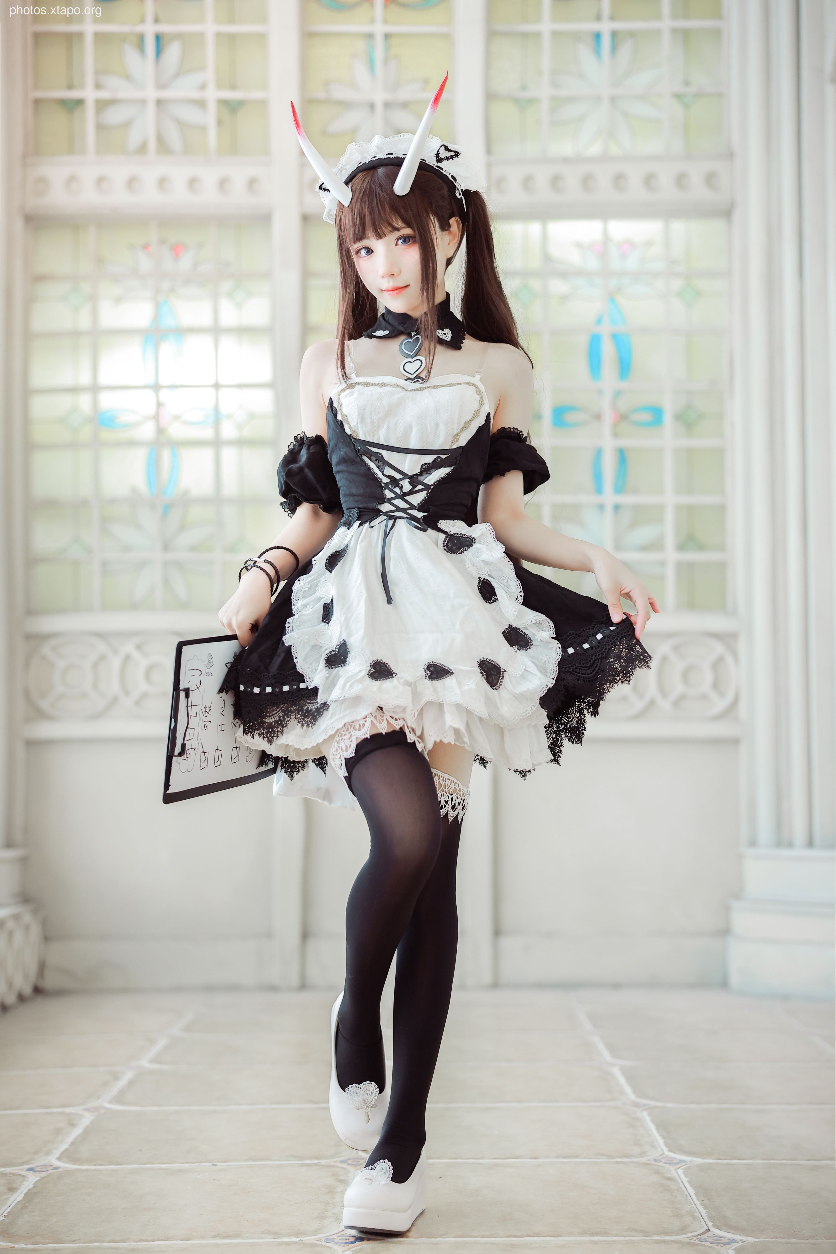 A cosplay portrait of a young woman dressed as a maid with devil horns, holding a clipboard. She is wearing a black and white maid dress, black thigh-high stockings, and white shoes.