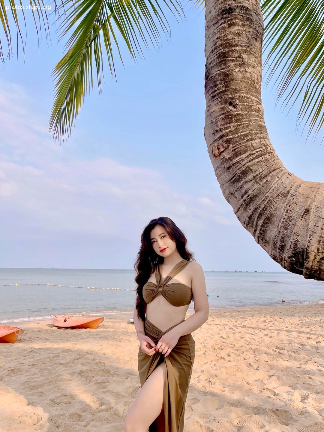 Woman in a brown bikini and matching skirt poses on a tropical beach under a palm tree.