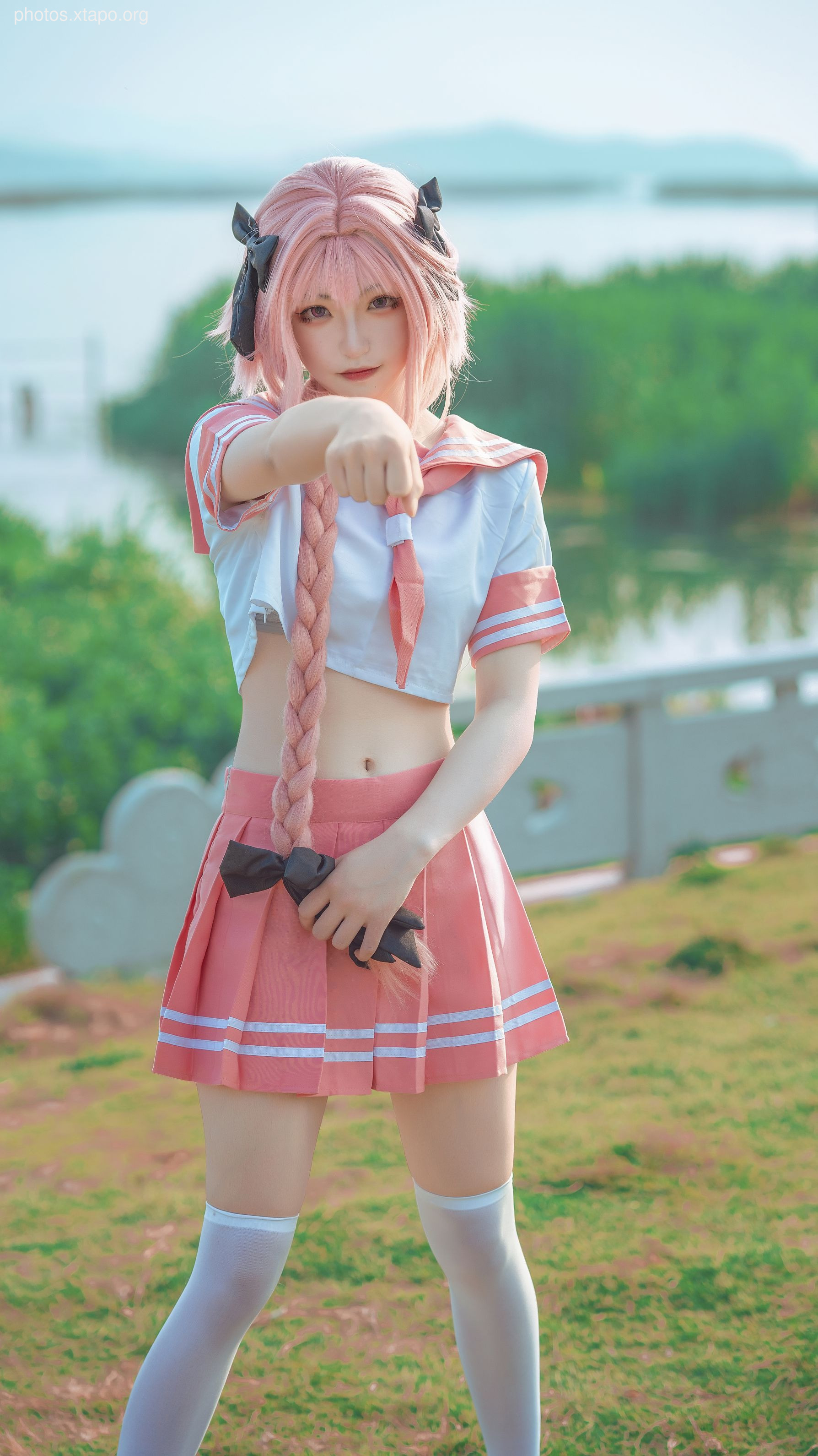 A cosplayer dressed as Astolfo from Fate/Grand Order, striking a playful pose in a picturesque outdoor setting.