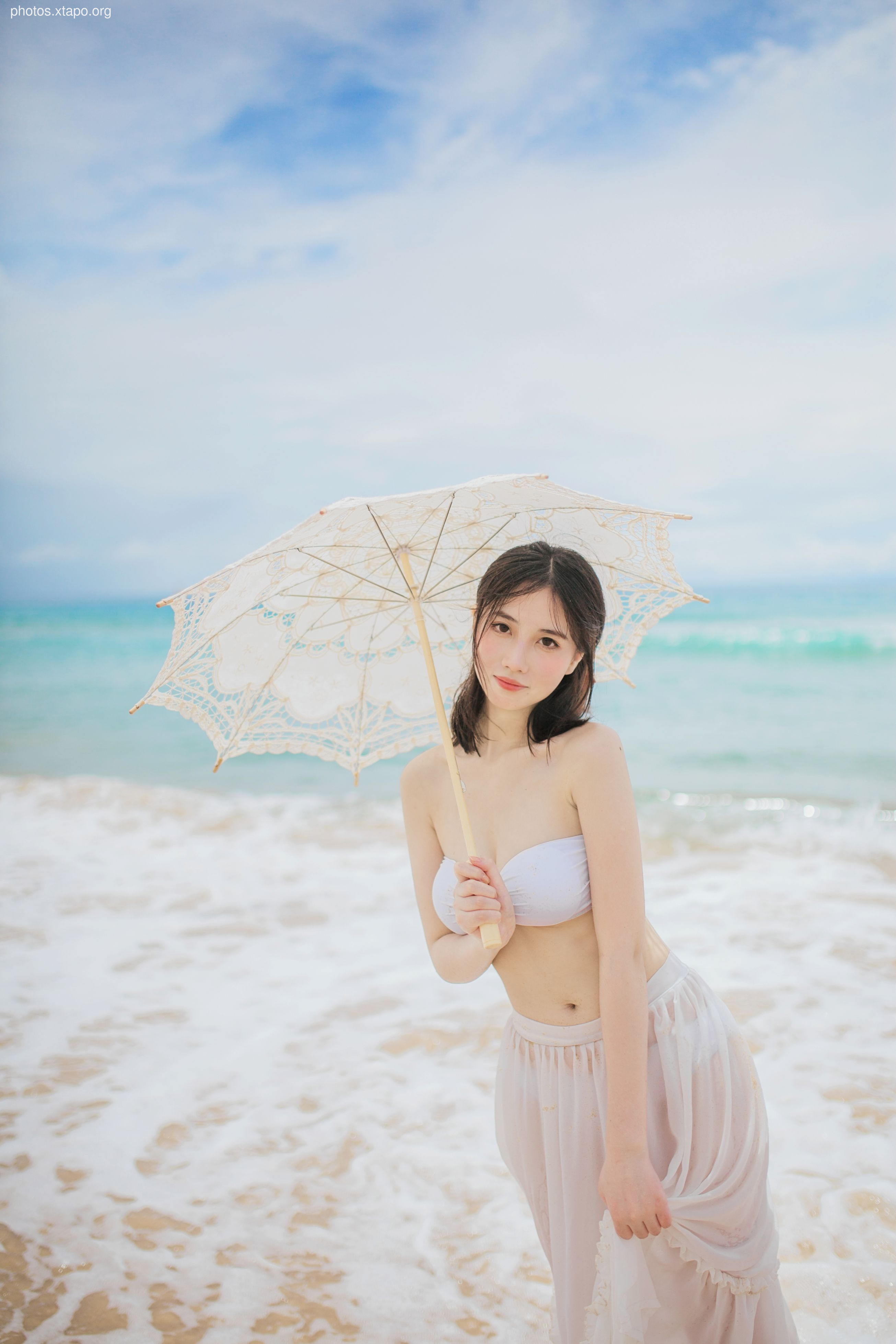 A woman in a white bikini and sheer skirt poses on a beach with a lace parasol.