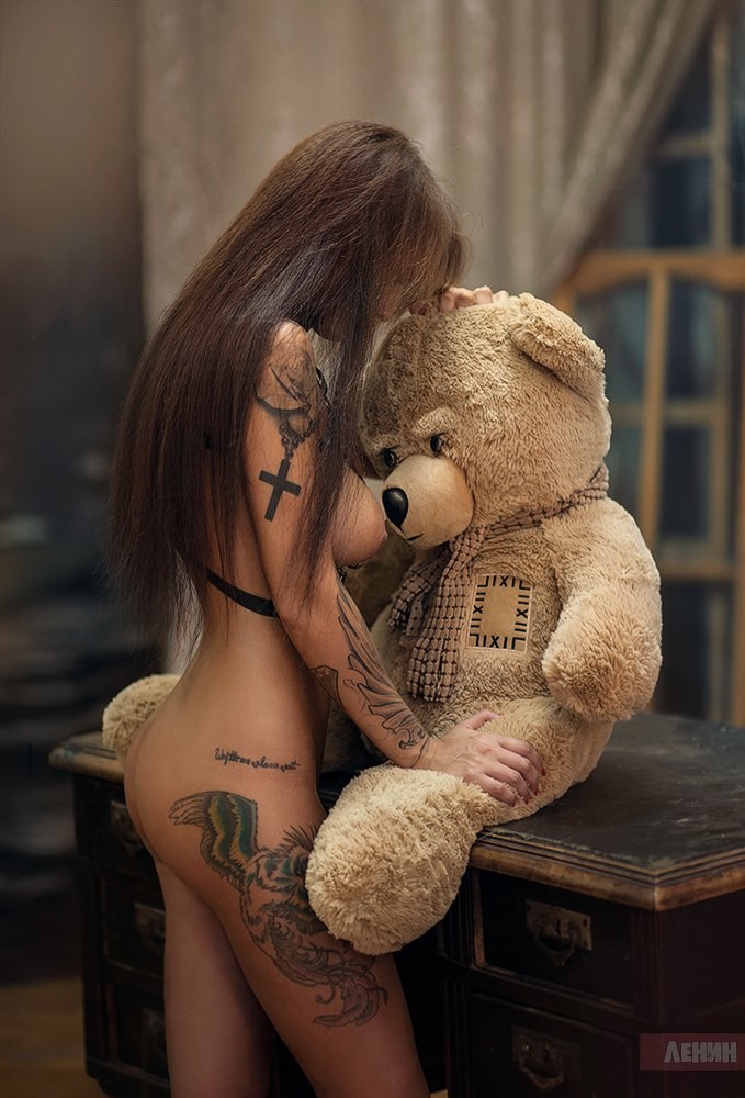 A woman with intricate tattoos embraces a large teddy bear, conveying a sense of comfort and vulnerability.