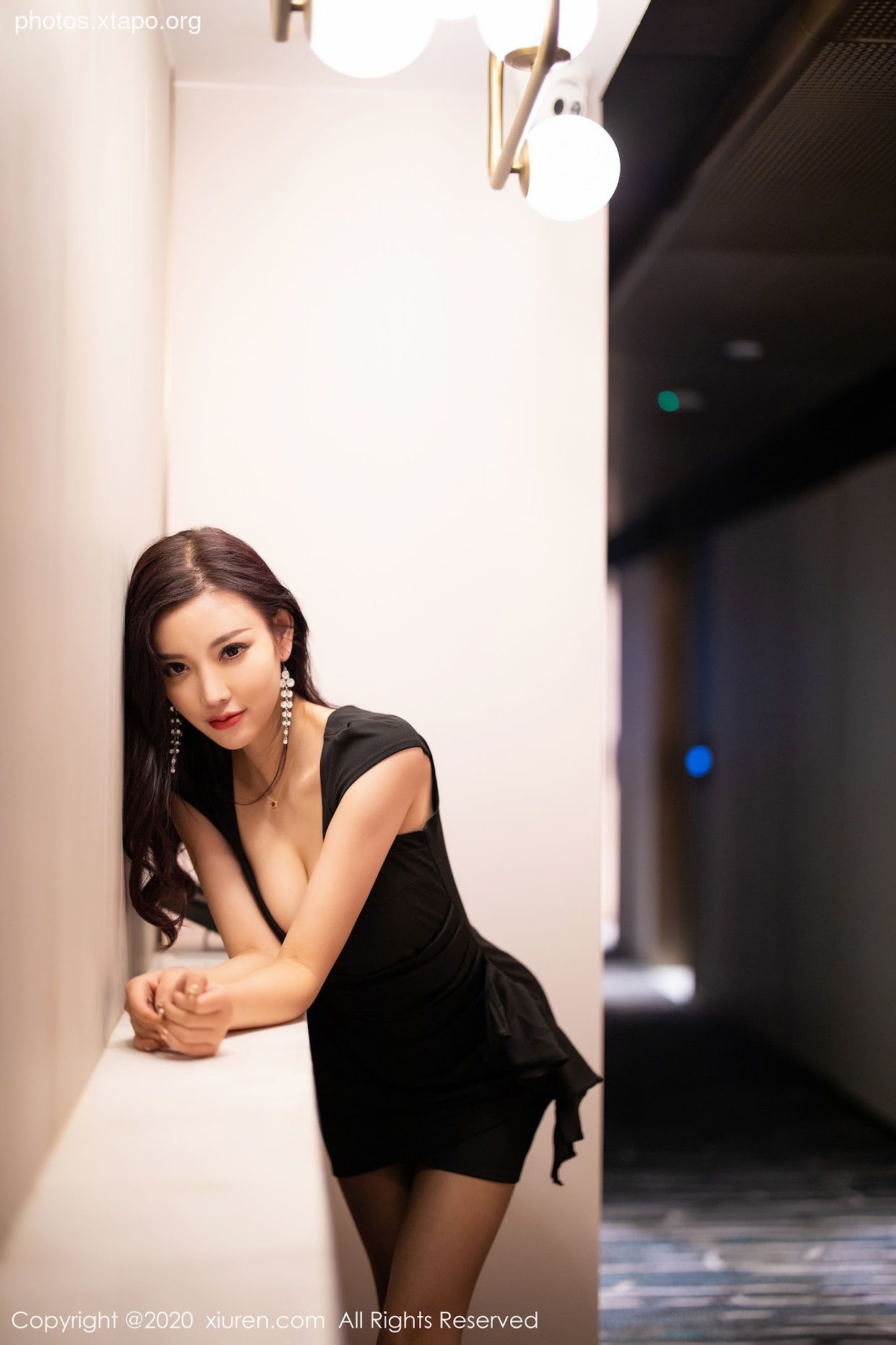 A woman in a little black dress poses against a wall in a dimly lit hallway.