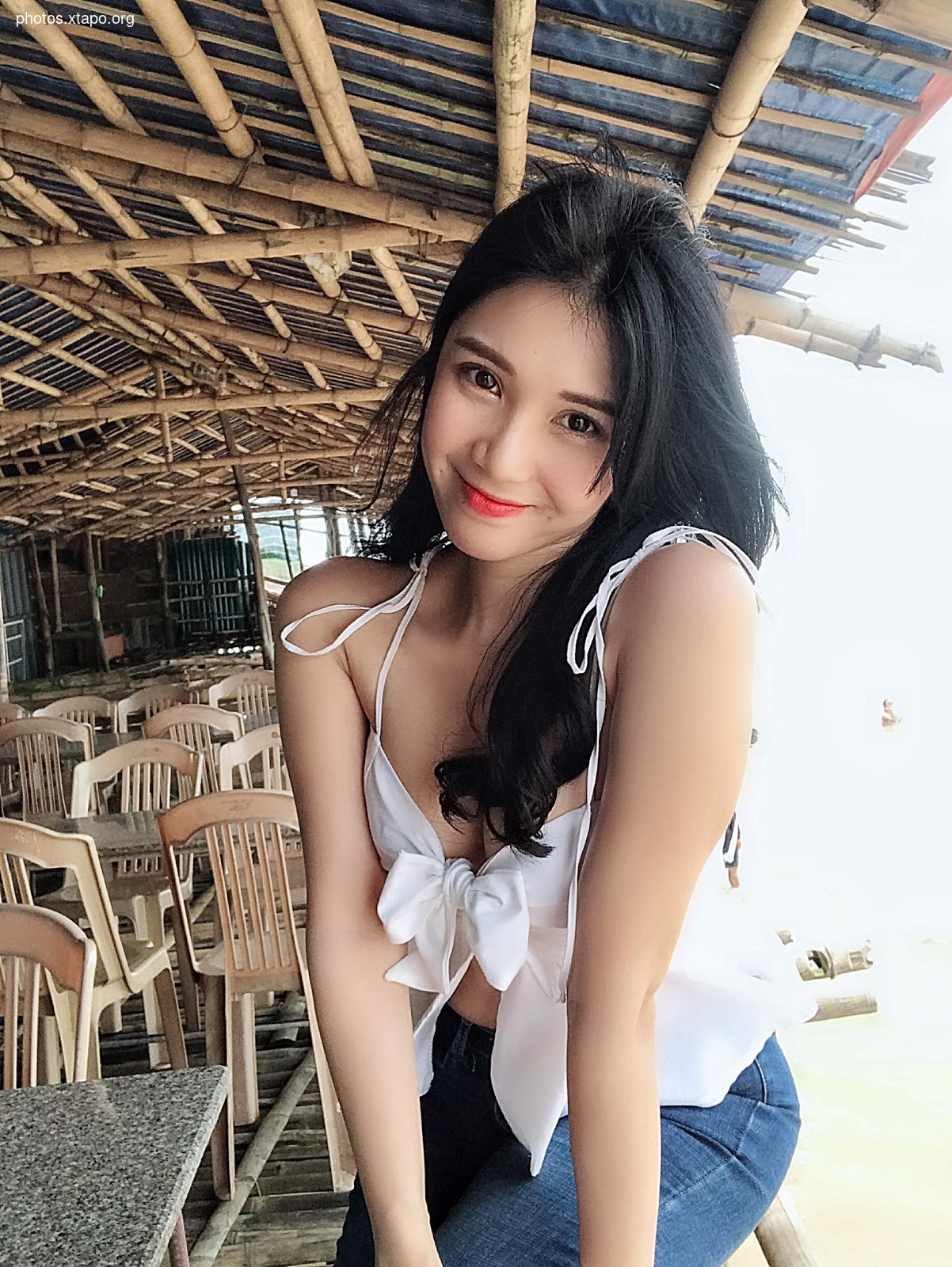 A young woman with long black hair smiles while wearing a white crop top and blue jeans. She is sitting under a bamboo structure.
