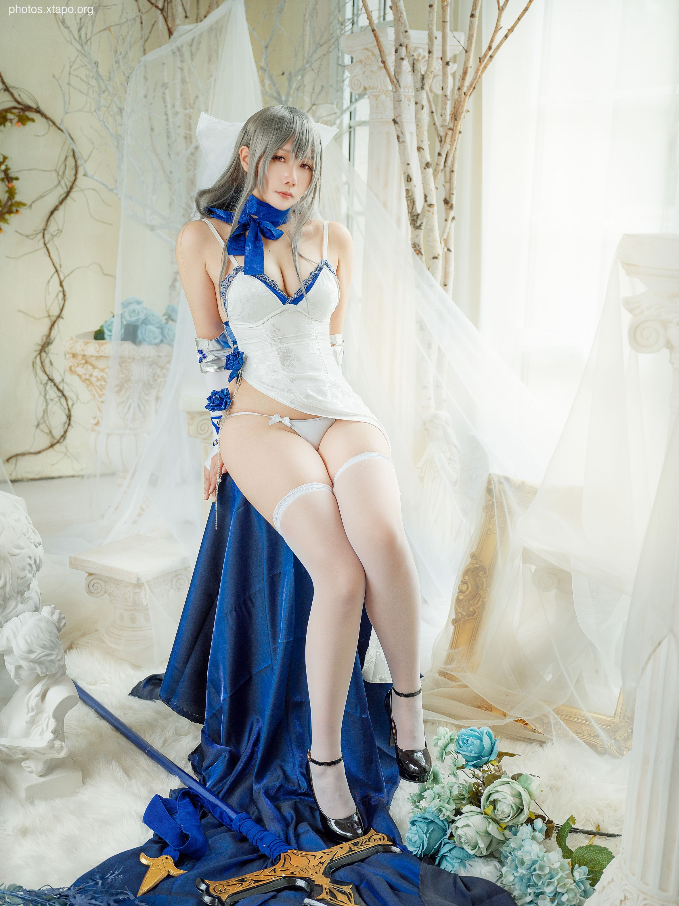 A stunning cosplay photoshoot featuring a model in a white and blue dress, striking a pose with a sword. The setting is elegant and dreamy.
