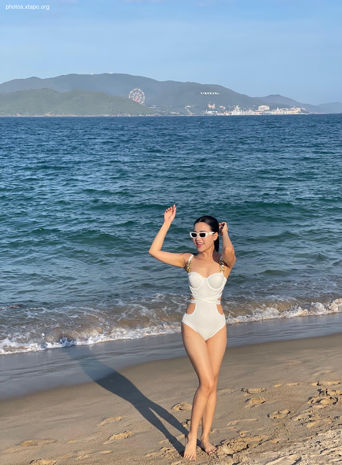A woman in a white swimsuit poses on a beach with a Ferris wheel and mountains in the background.