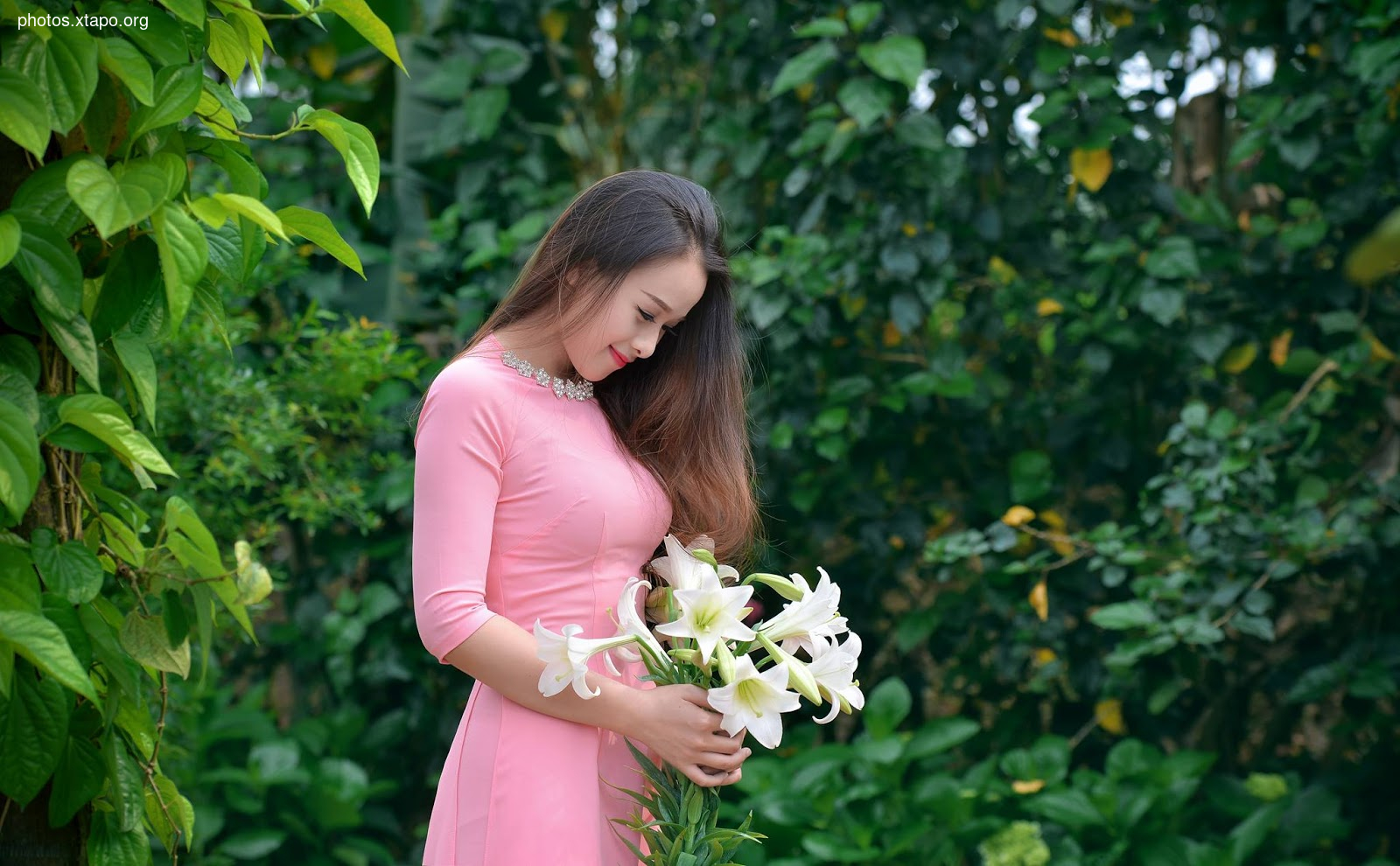 A young woman in a pink dress stands in a garden, gently holding a bouquet of white lilies.  The lush green foliage provides a vibrant backdrop.