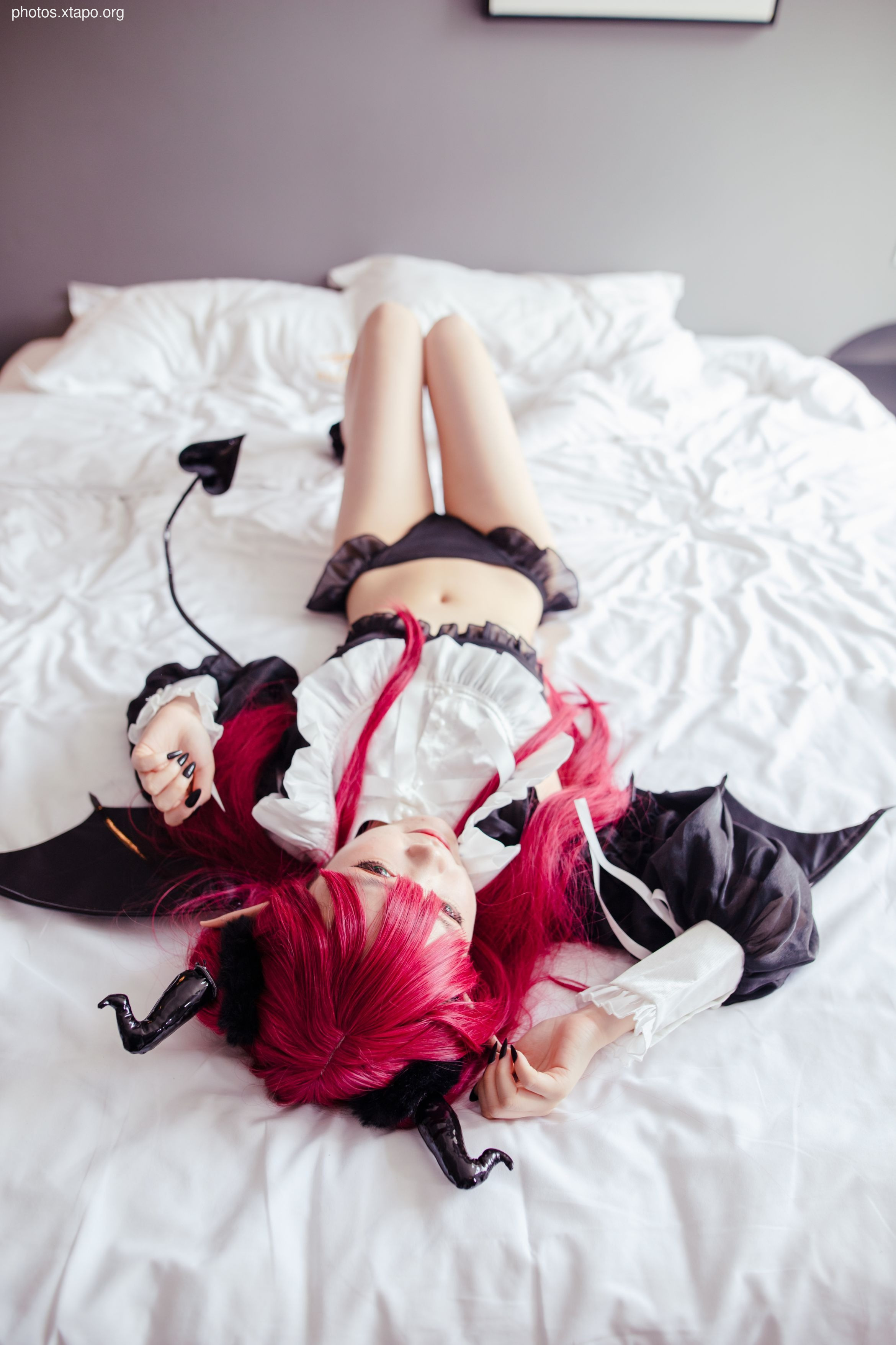 A young woman with vibrant red hair and devil horns lies on a bed, dressed in a black and white lingerie-inspired outfit.  She is playfully posing for the camera.