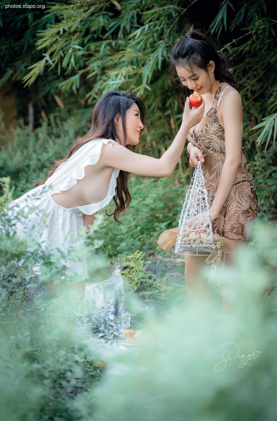 Two young women in summer dresses share a red apple in a lush green garden. One woman wears a white dress, the other a floral-patterned dress. They appear happy and carefree.