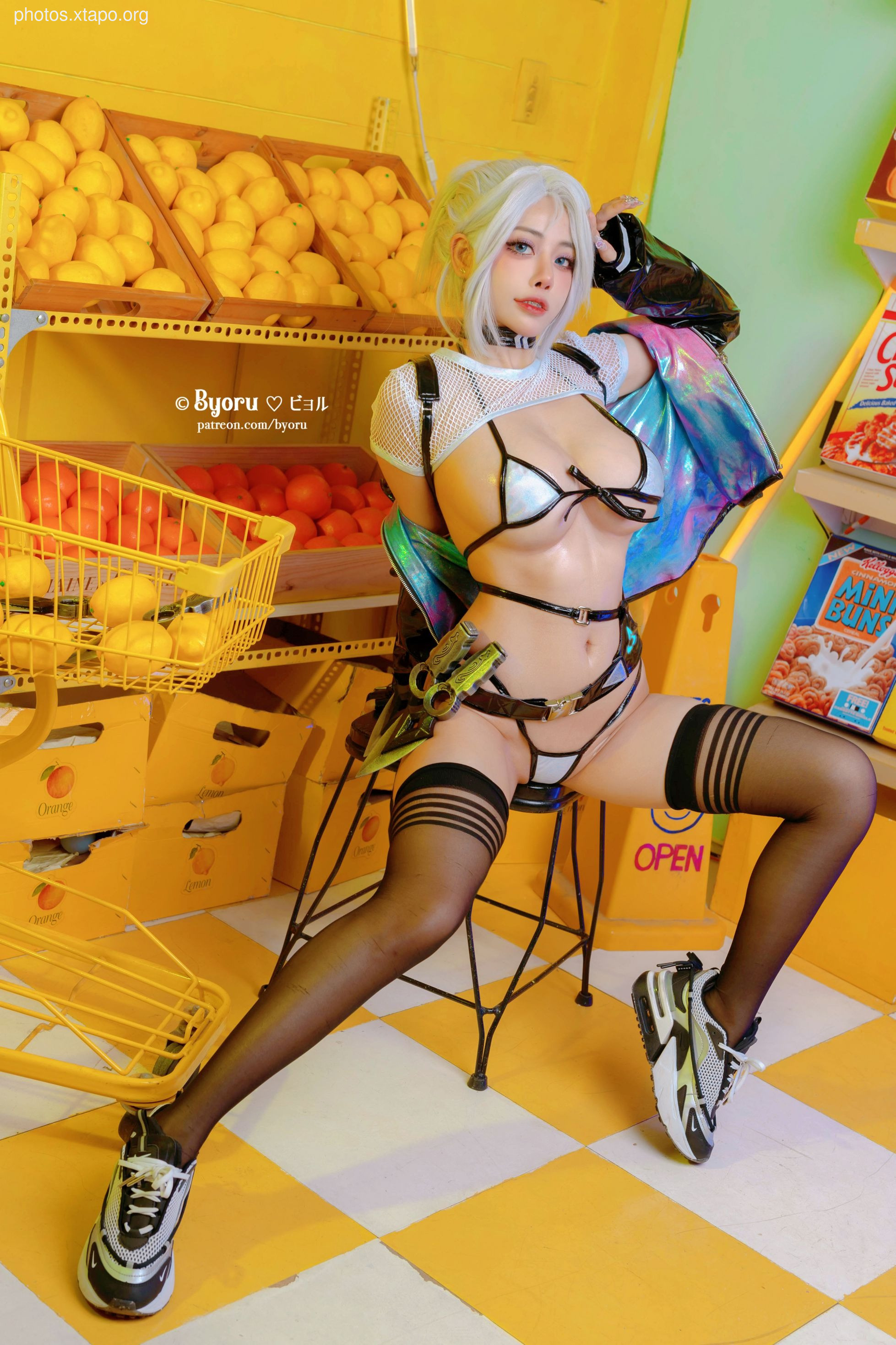 A model in a cyberpunk-inspired cosplay poses in a brightly colored store filled with fruit. She is wearing a white and silver bikini top, black stockings, and sneakers.