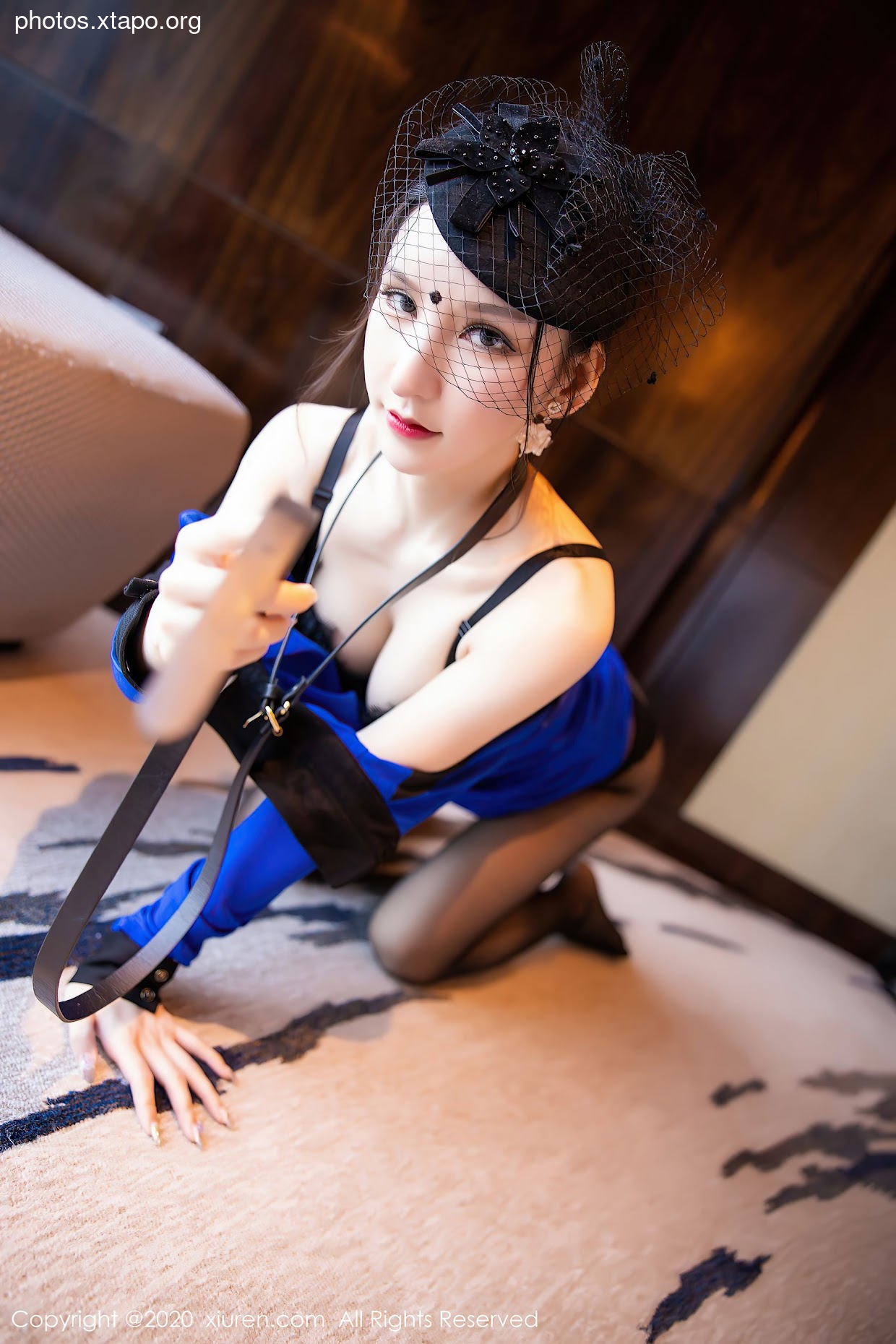 A woman wearing a blue dress and a black hat with a veil is holding a whip. She is kneeling on a carpeted floor.