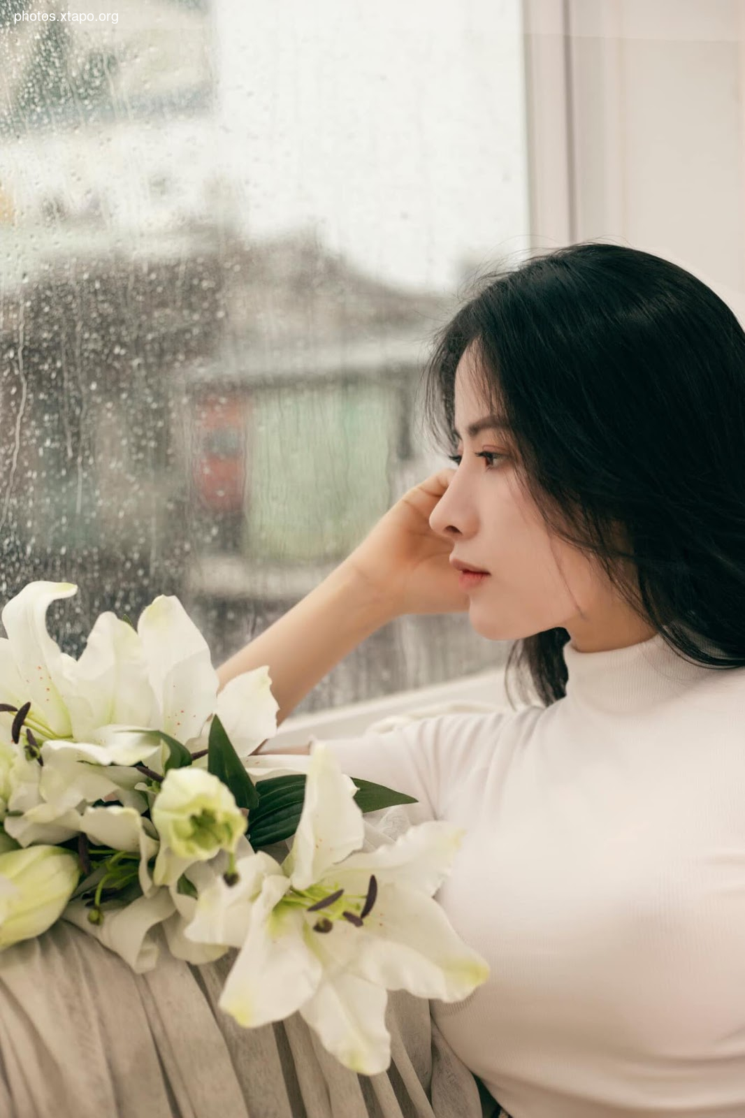 A young woman sits by a rain-streaked window, lost in thought. White lilies rest on her lap, adding to the serene yet melancholic mood.