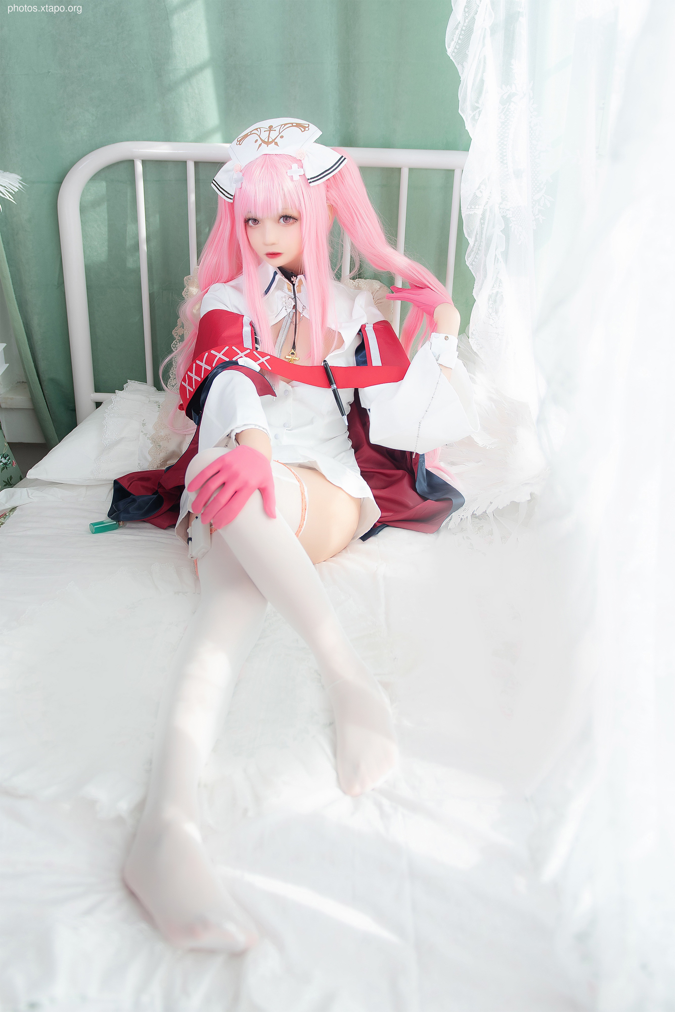 A model poses in a sailor-themed cosplay outfit, featuring a white and red dress with pink hair. The setting is a bedroom with white bedding and curtains.