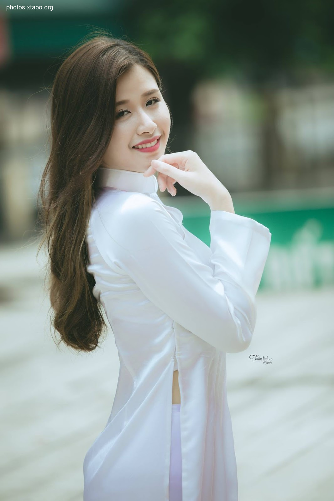 A young woman smiles gently, her long brown hair cascading down her shoulders as she stands in a pristine white Ao Dai. The soft focus background enhances her serene beauty.