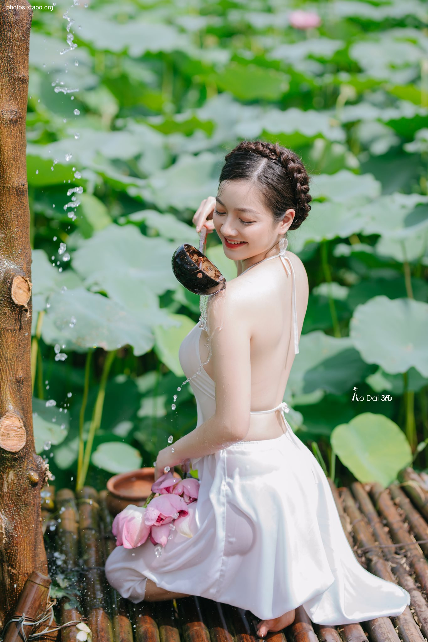 A young woman in a white dress bathes in a tranquil garden filled with lush green lotus plants.  She uses a coconut shell to scoop water as it flows past her.