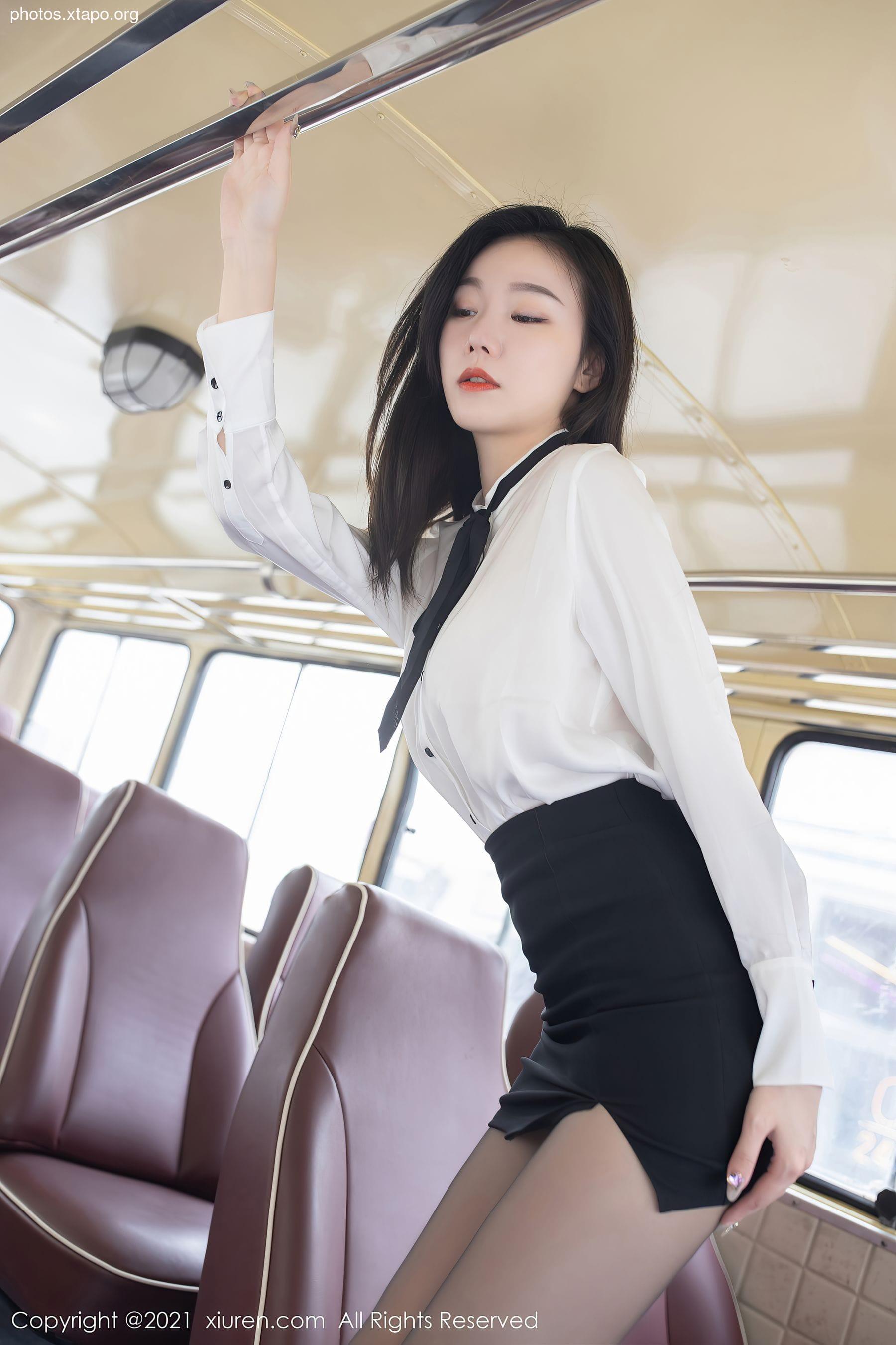 A young woman in a white shirt and black skirt stands on a public bus, holding onto a handrail.