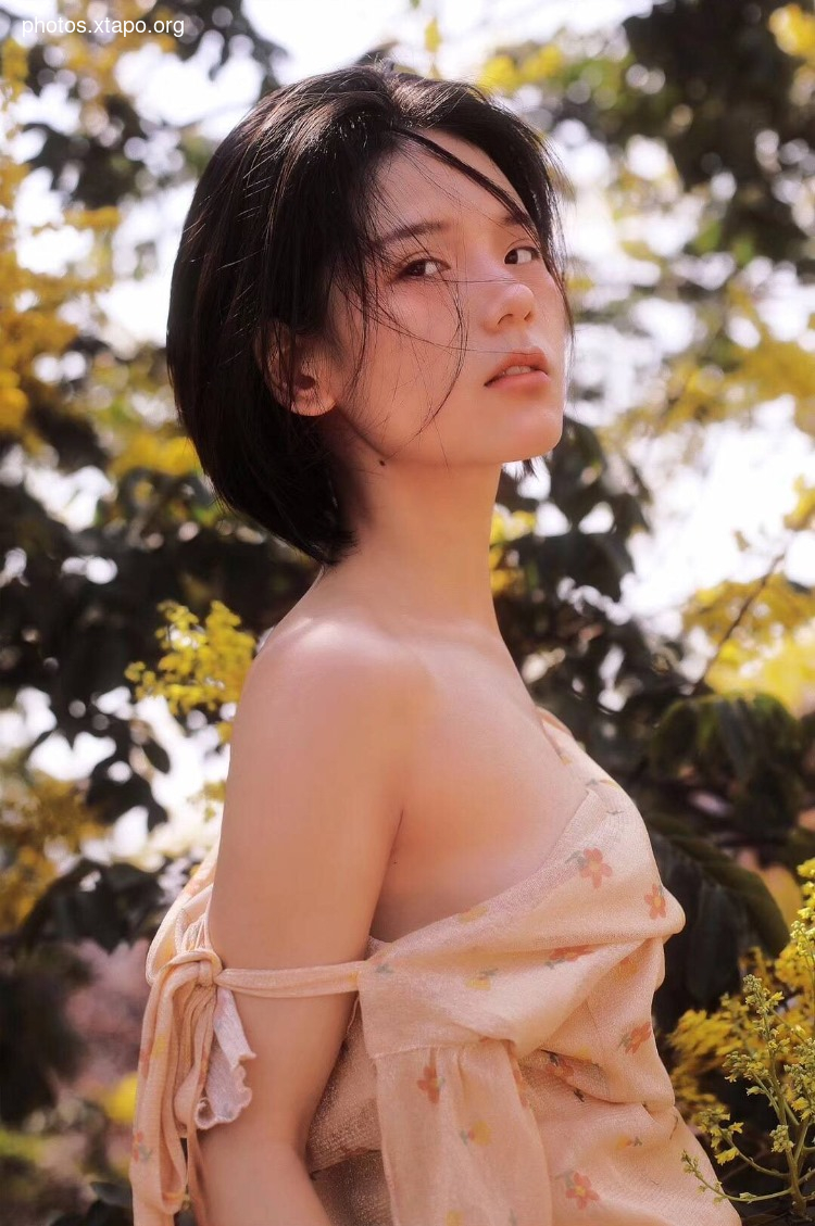 A young woman with short black hair stands in a field of yellow flowers, wearing a peach-colored dress with floral print.
