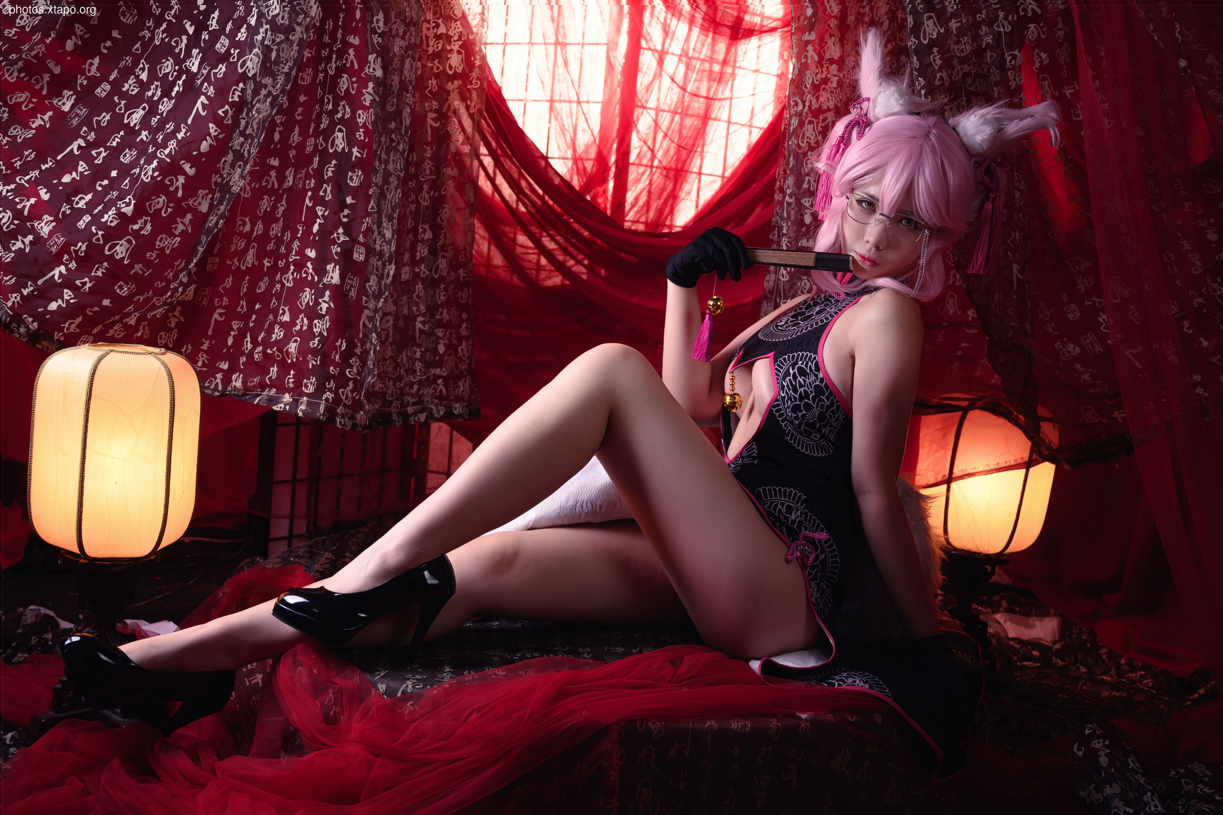A model in cosplay, dressed in a traditional Chinese-style black and white cheongsam with pink fox ears, holds a decorative fan. The setting is dimly lit, with red fabrics and lanterns creating a dramatic atmosphere.
