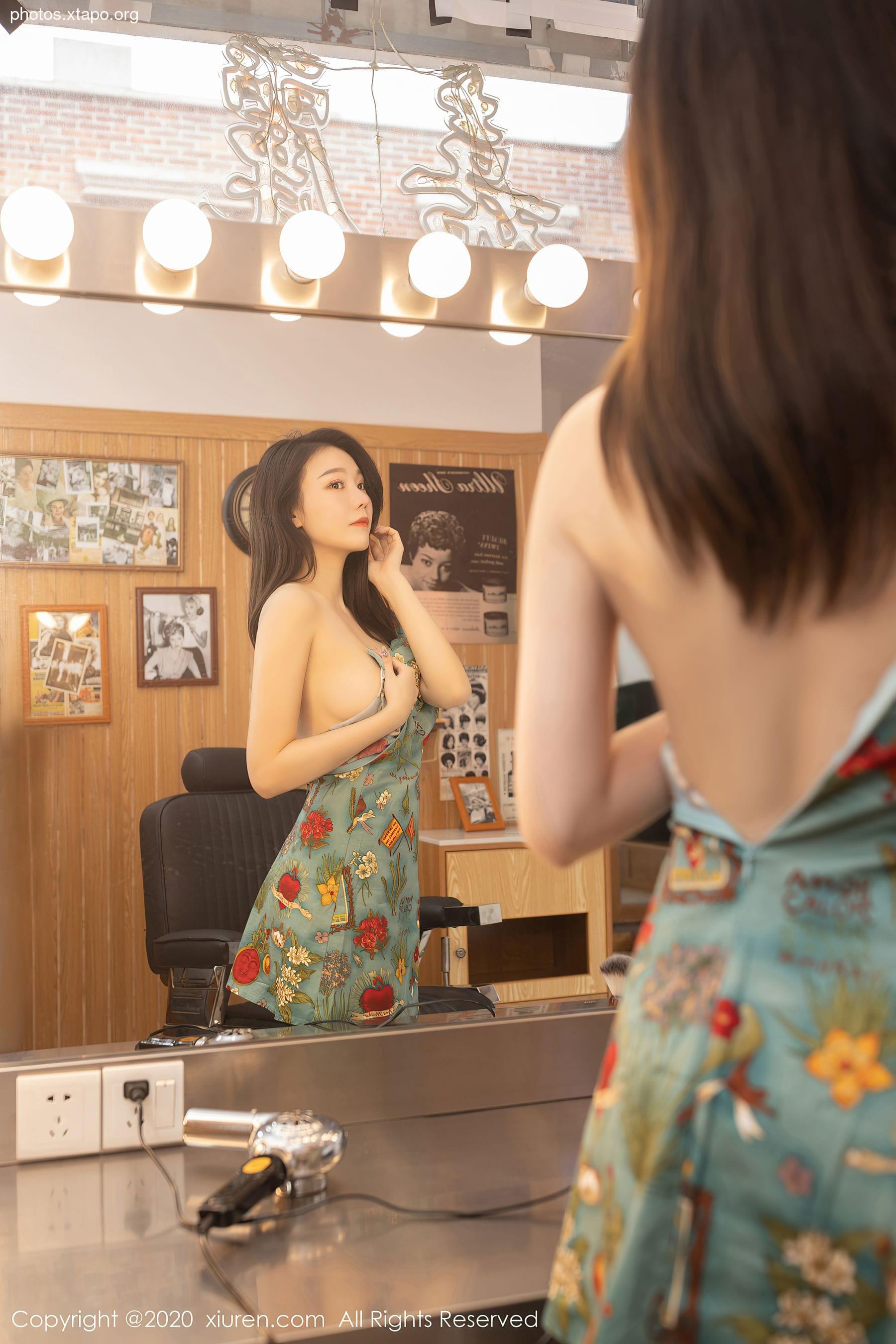 A young woman in a floral dress stands before a mirror, admiring her reflection. The setting appears to be a vintage-style barbershop.