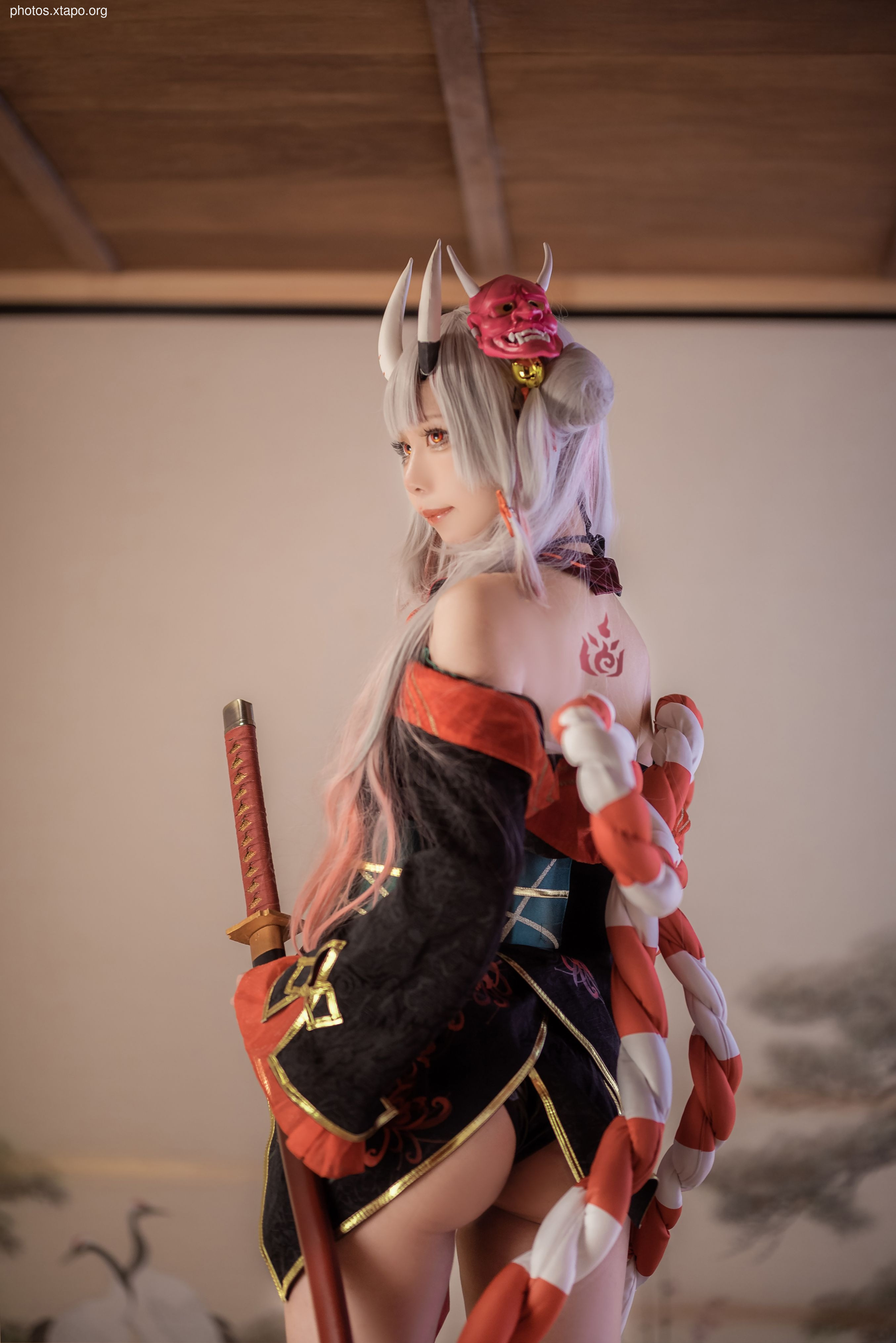 A captivating cosplay of a female warrior, adorned in intricate Japanese attire and wielding a katana. Her costume features a striking red and black color scheme, complemented by a traditional oni mask in her hair.  The image is well-lit, showcasing fine details and textures.