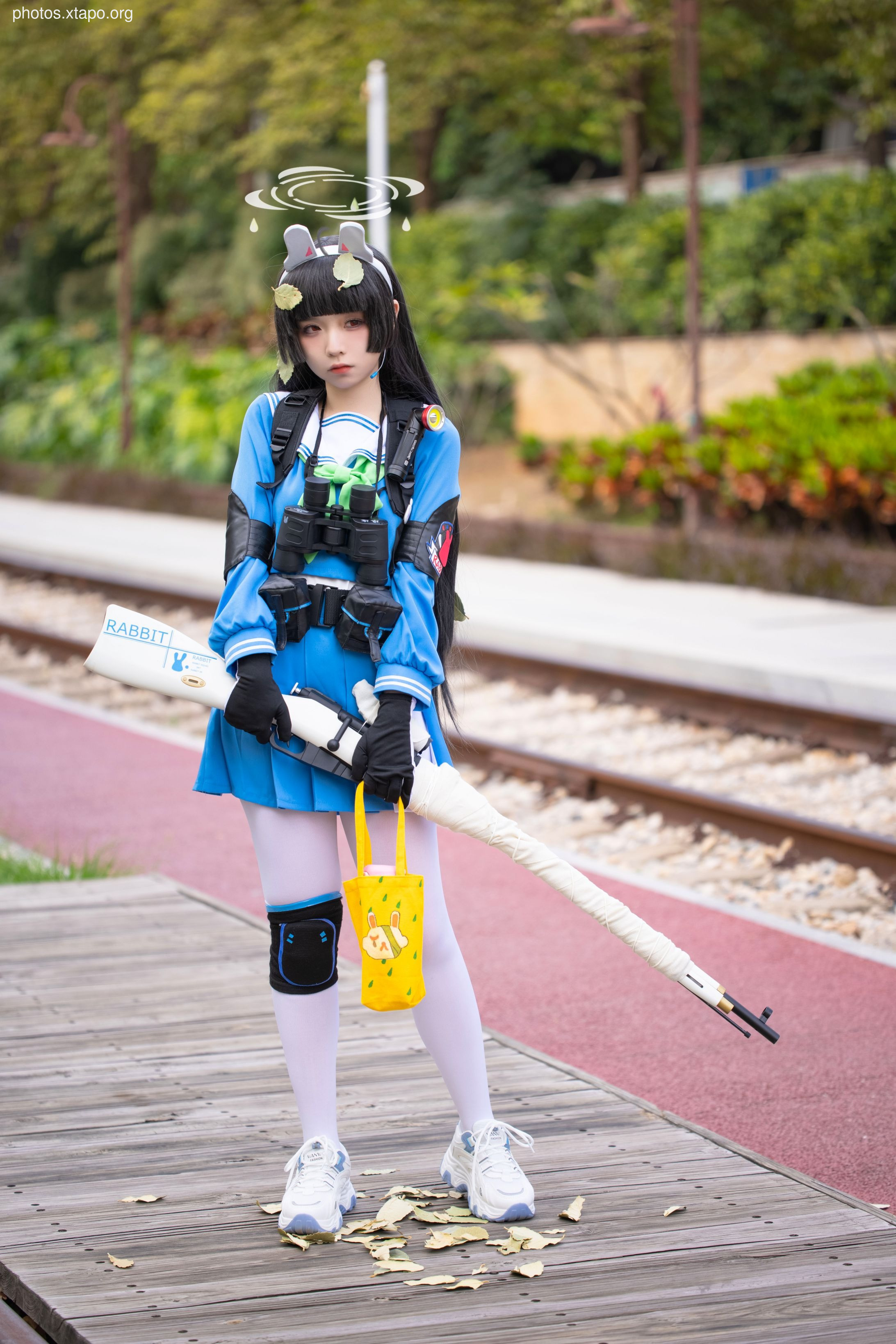 Cosplay of a girl in a blue and white outfit holding a rifle, standing on a railway platform.