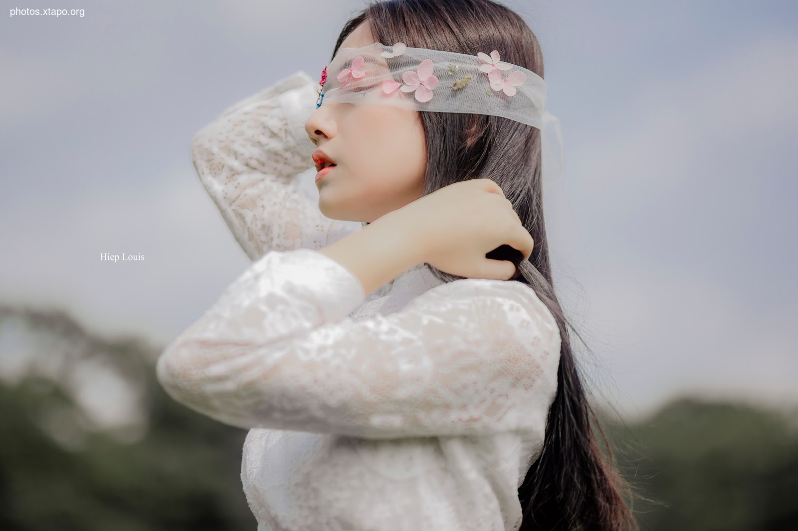 A young woman in a white lace dress, her eyes covered by a delicate floral blindfold, gazes pensively into the distance.  The soft lighting and serene background enhance the ethereal mood of the image.