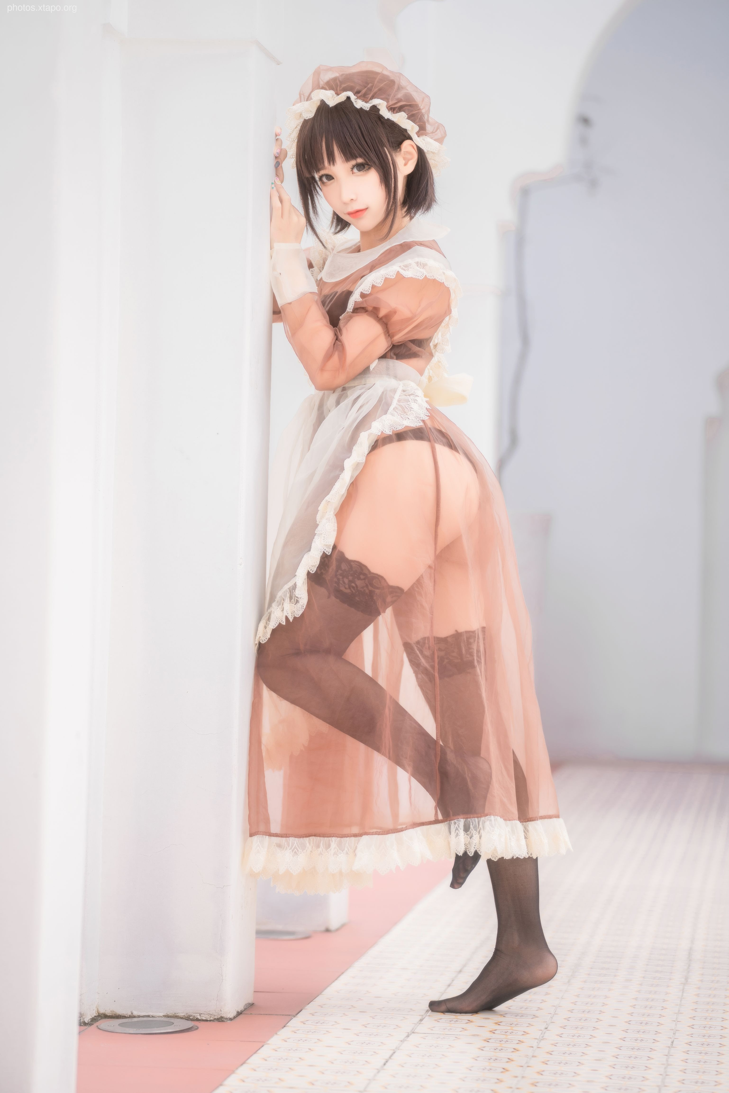 A playful maid in a sheer brown dress leans against a white pillar.