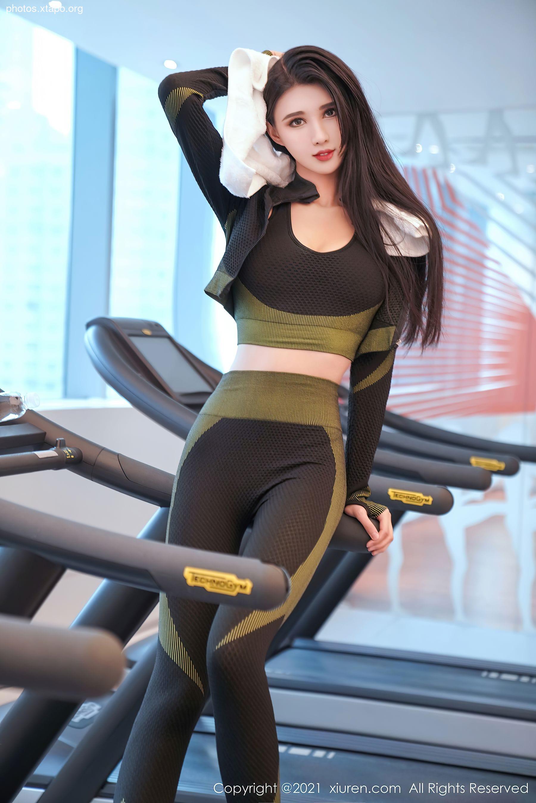 A woman in athletic wear wipes her forehead with a towel while standing on a treadmill in a gym.