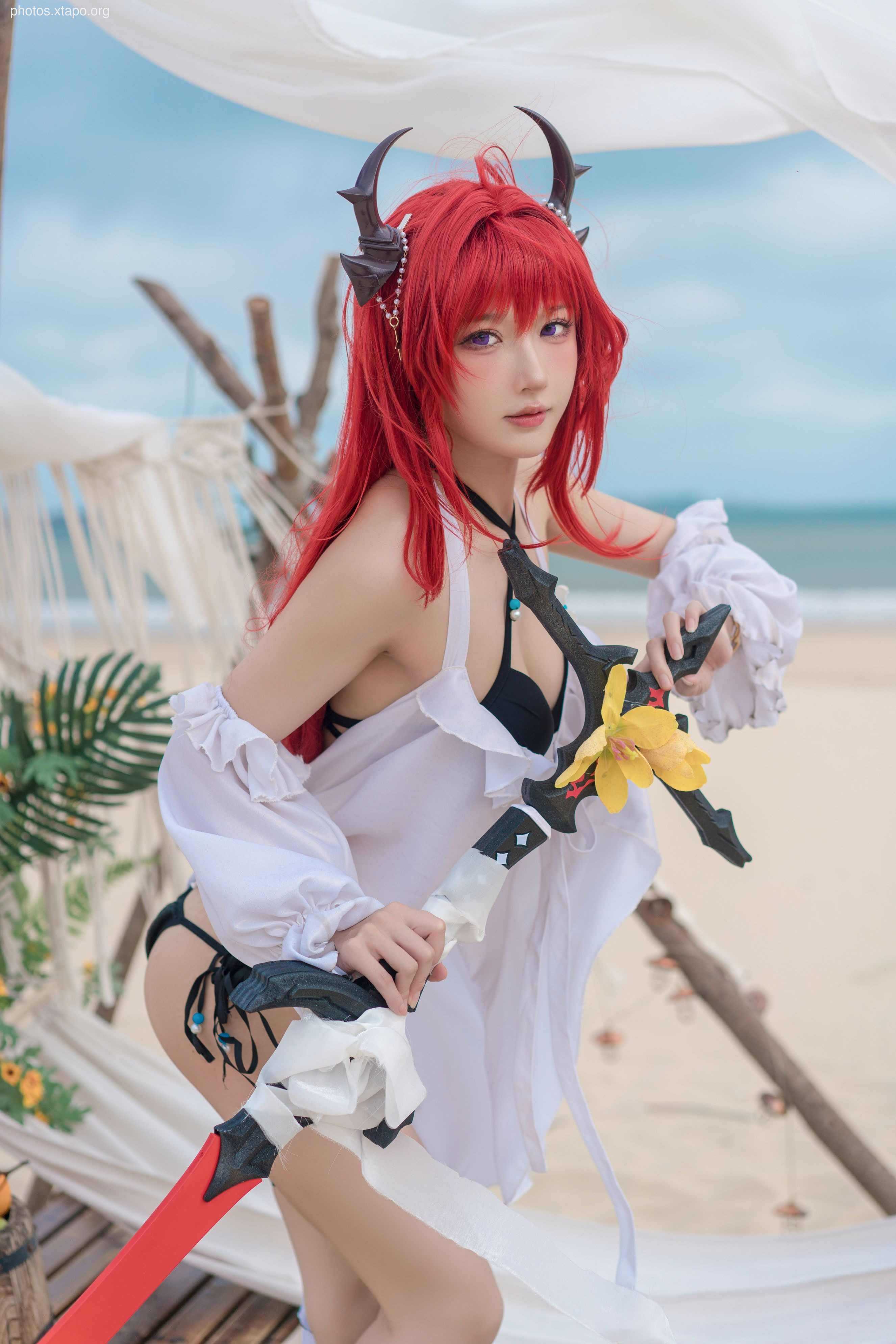 A cosplayer, dressed as a character with red hair and devil horns, poses on a beach with a large sword.  She wears a white shirt and bikini bottoms.
