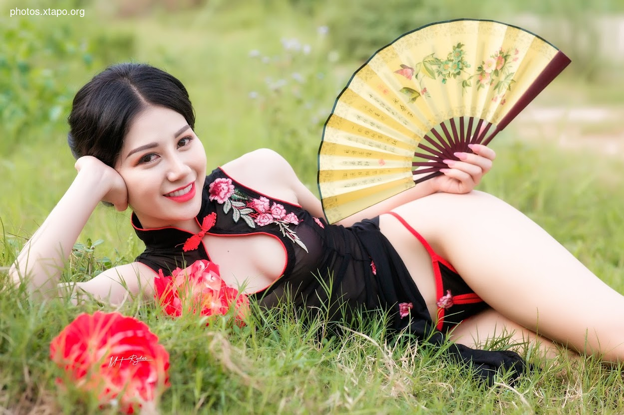 A young woman in a black cheongsam with red floral embroidery lies in a grassy field, holding a traditional Chinese fan.  She is wearing red lingerie. 