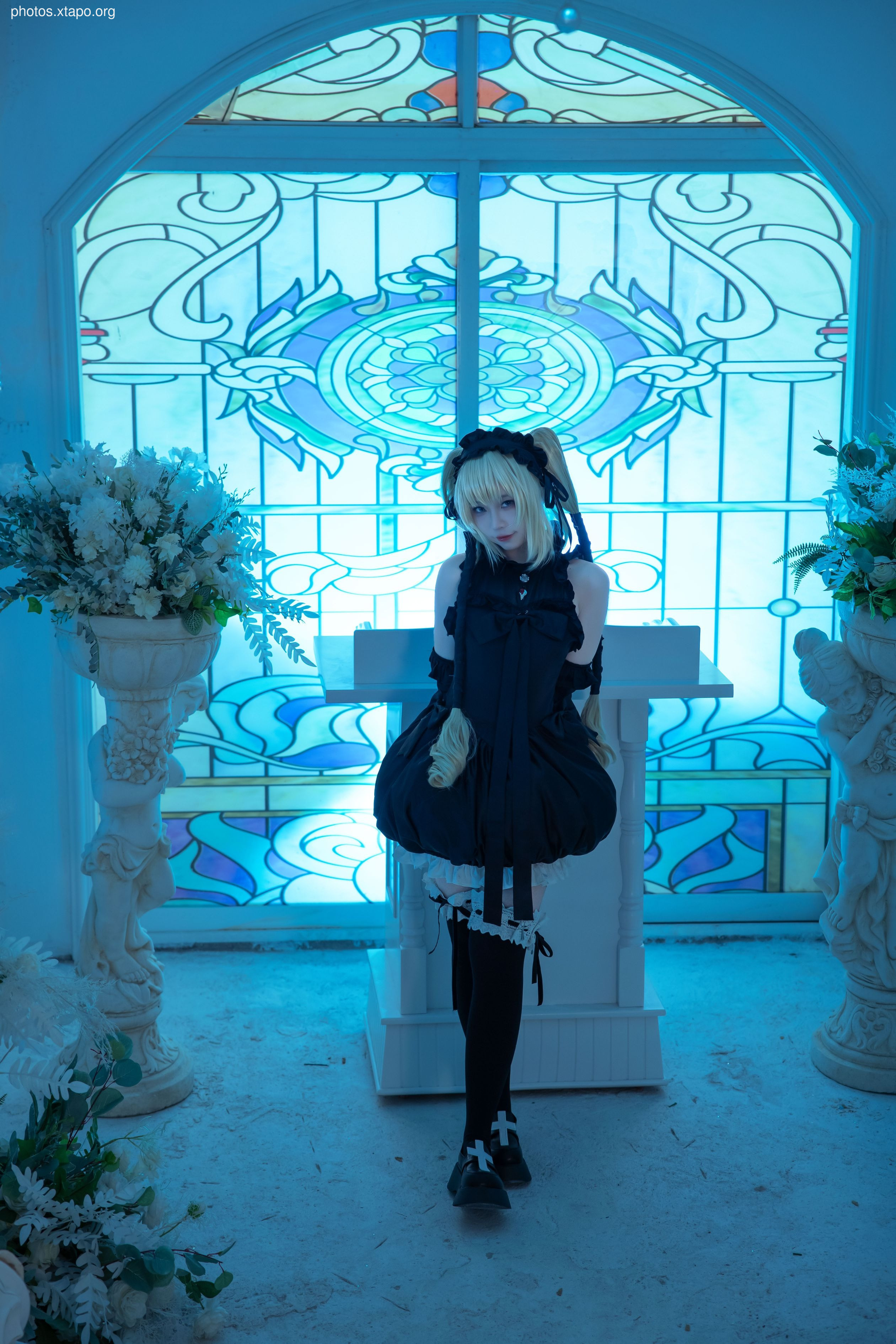 A cosplayer dressed as a gothic lolita stands in a chapel with blue stained-glass windows. She is wearing a black dress with white lace trim and black tights.