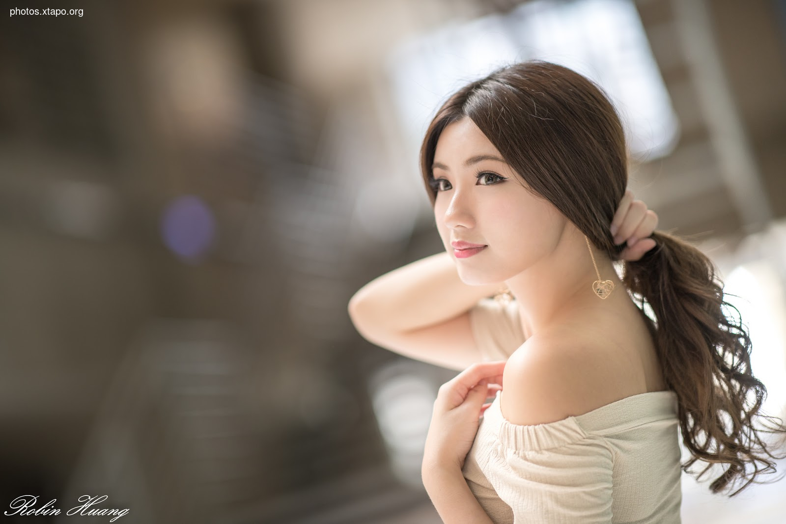 Portrait of a young woman with long brown hair, wearing a light beige off-shoulder top. She is looking to the side and slightly smiling. The background is blurred and out of focus.