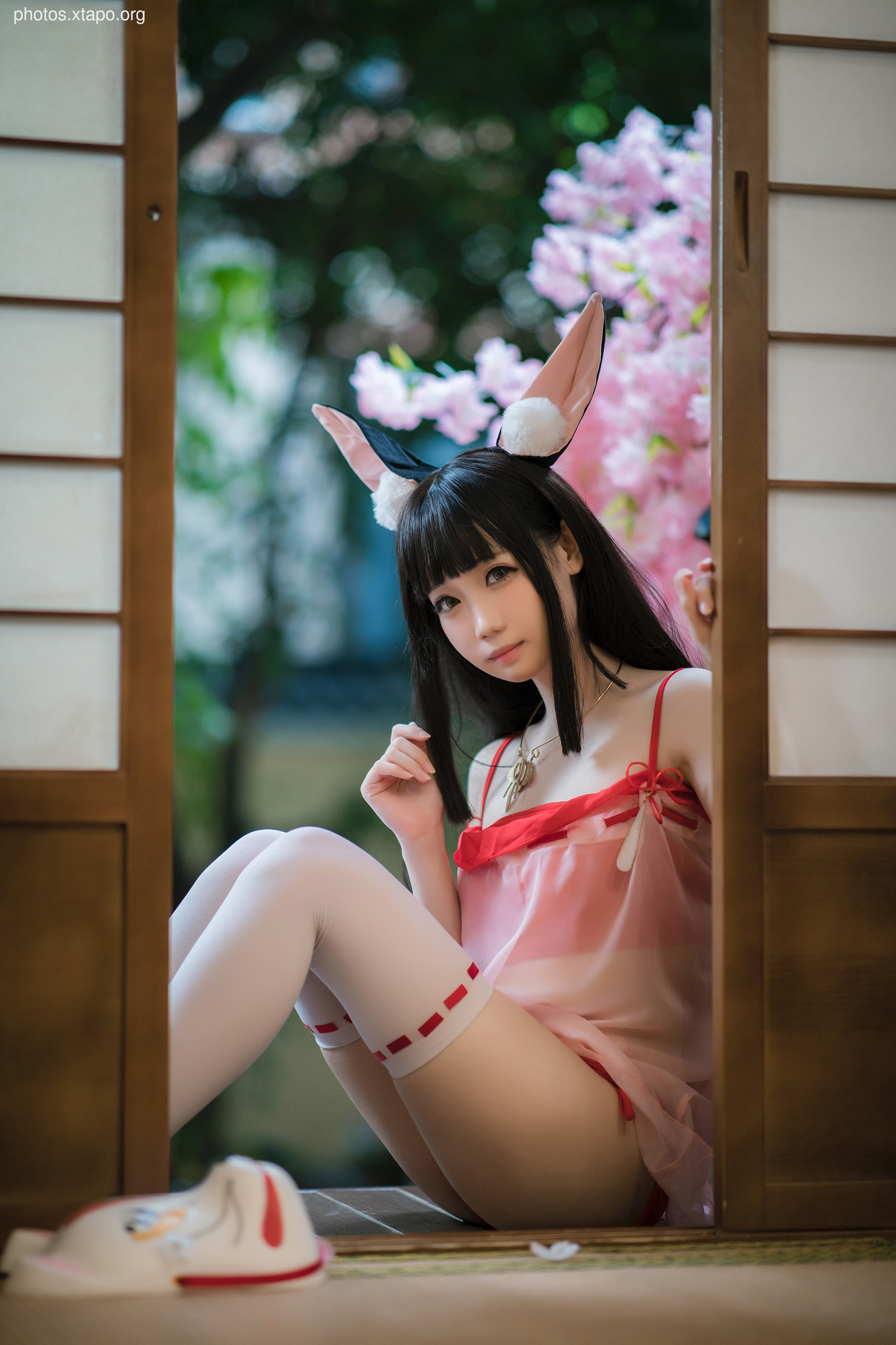 A young woman in a bunny girl cosplay sits near a Japanese-style screen.  She is wearing a pink outfit and white thigh-high socks with red accents.