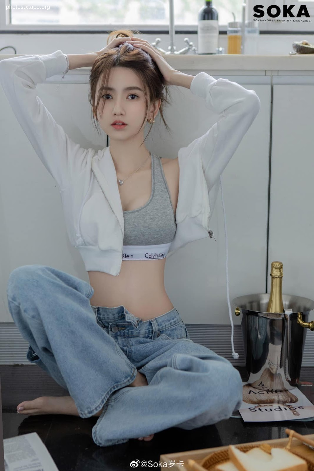 A young woman poses in a white hoodie, gray sports bra, and light blue jeans. The image is set against a minimalist kitchen backdrop.