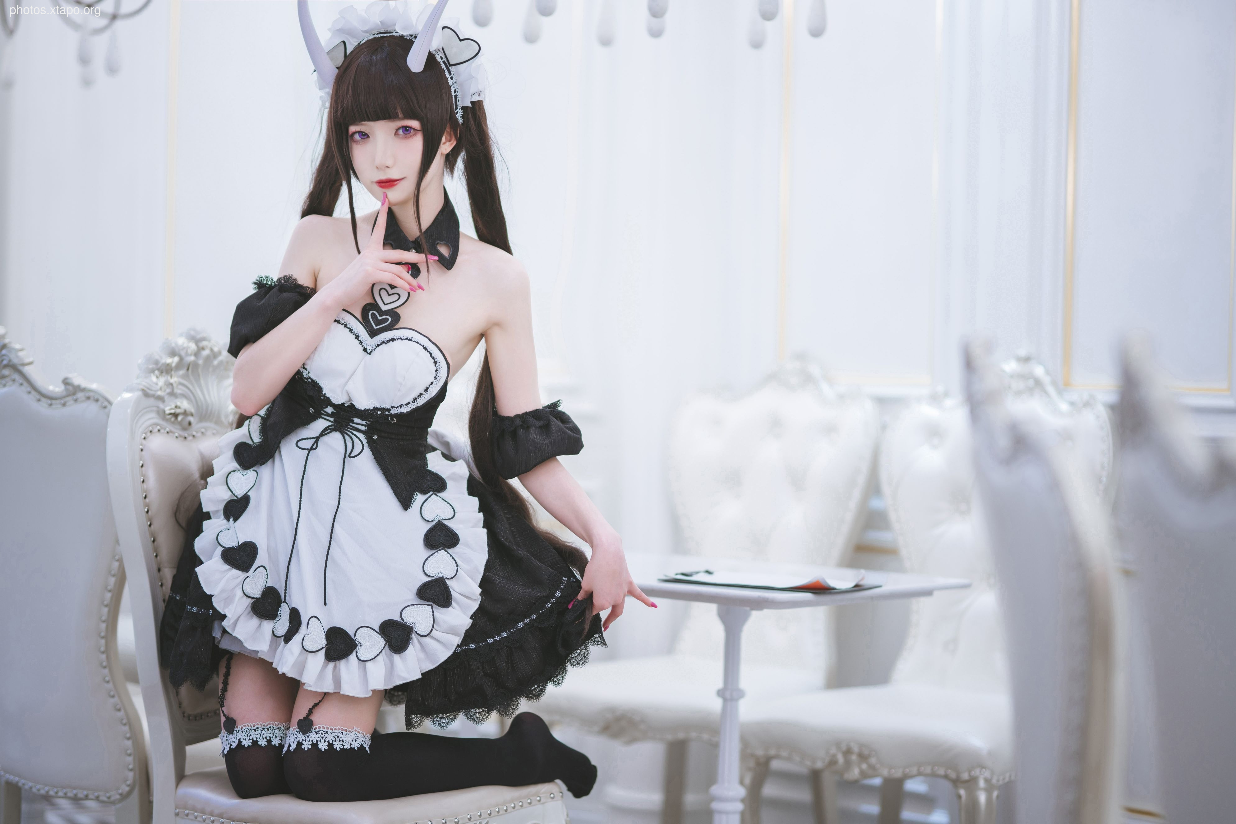 A young woman dressed as a devilish maid poses for a photograph. She is wearing a black and white maid outfit with heart-shaped details and devil horns. She has long black hair and is giving a mischievous expression.