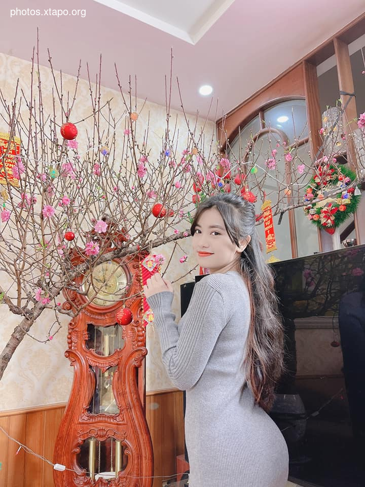 A young woman in a stylish gray dress poses gracefully beside a beautifully decorated peach blossom tree, creating a charming and festive atmosphere.