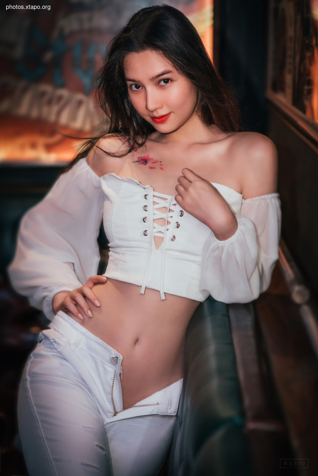 A captivating portrait of a young woman with striking features and a delicate floral tattoo. She is dressed in a stylish off-the-shoulder top and white pants, exuding elegance and charm.