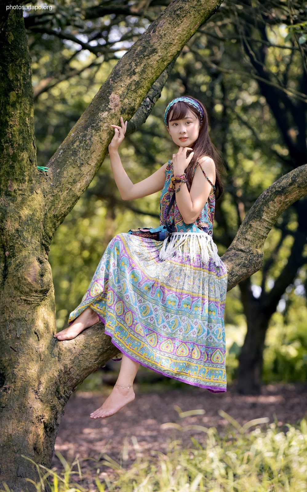 A young woman in a bohemian-style dress sits on a tree branch in a lush forest.  Her relaxed pose and colorful clothing create a peaceful and serene image.