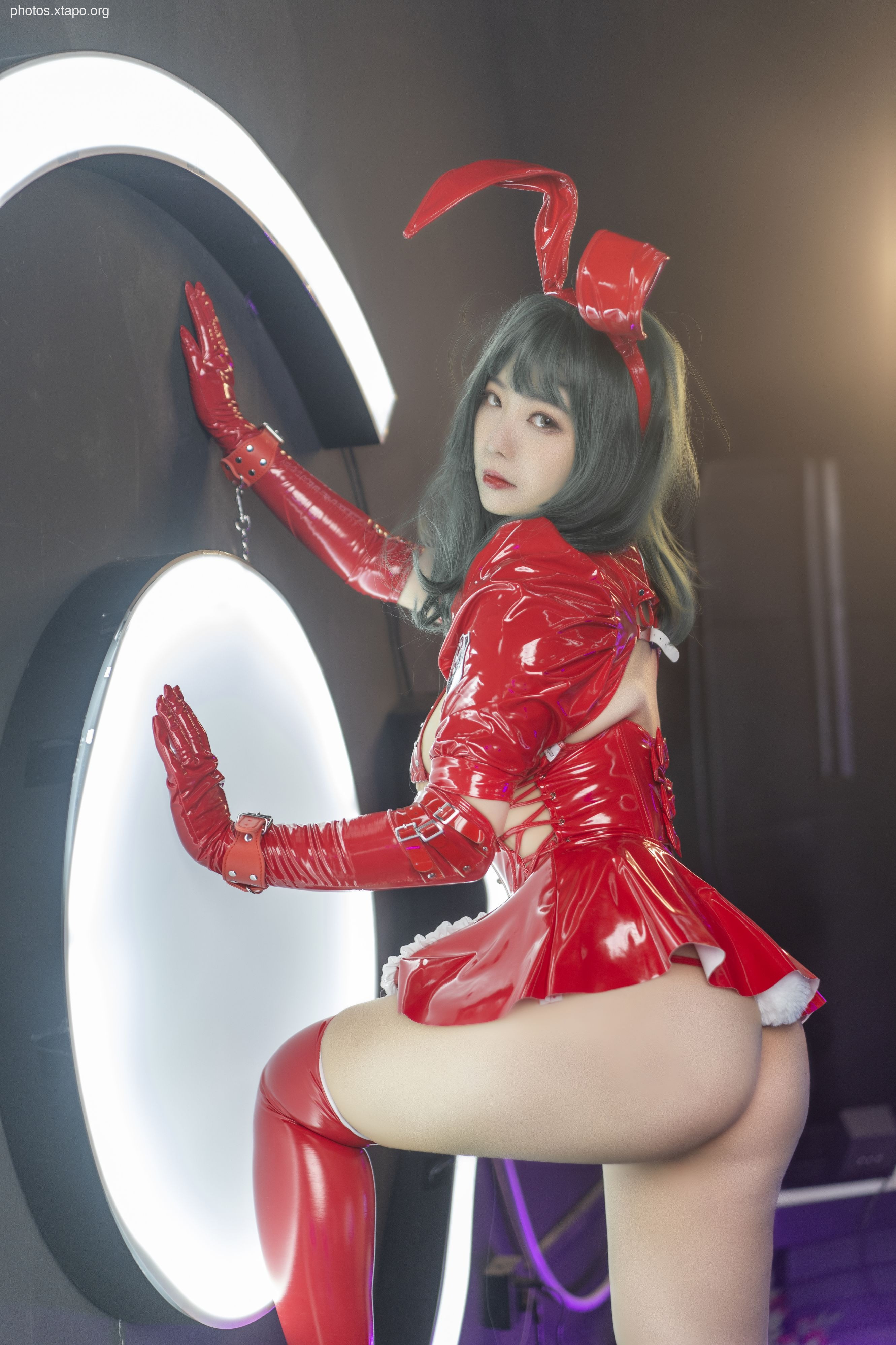 A woman in a red latex bunny girl costume poses against a futuristic backdrop.  She is wearing gloves and cuffs, suggesting a playful or mischievous theme.