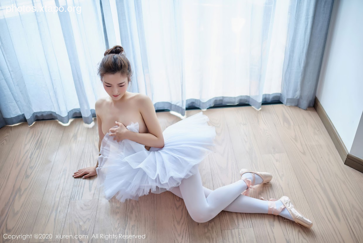 A young woman in a white tutu sits on a wooden floor, adjusting her clothing. She is wearing ballet pointe shoes and has her hair pulled back in a bun. The background is blurred but shows a bright window.