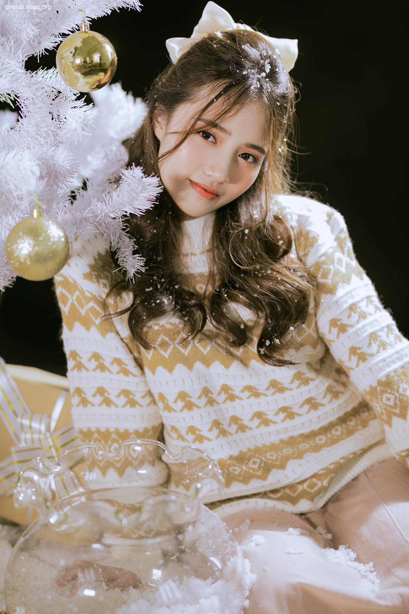 A young woman in a festive sweater poses by a Christmas tree.