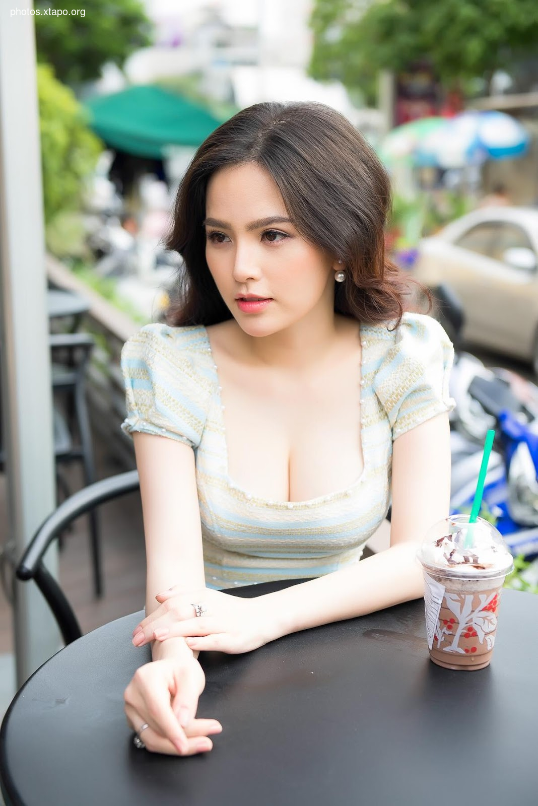 A young woman with long dark hair sits at an outdoor table, enjoying a coffee drink. She is wearing a light-colored, short-sleeved top.