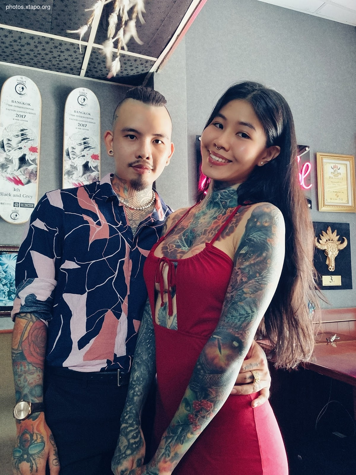 A tattooed couple poses for a photo. The woman is wearing a red dress and has long dark hair. The man is wearing a patterned shirt and has short dark hair. They are both smiling.