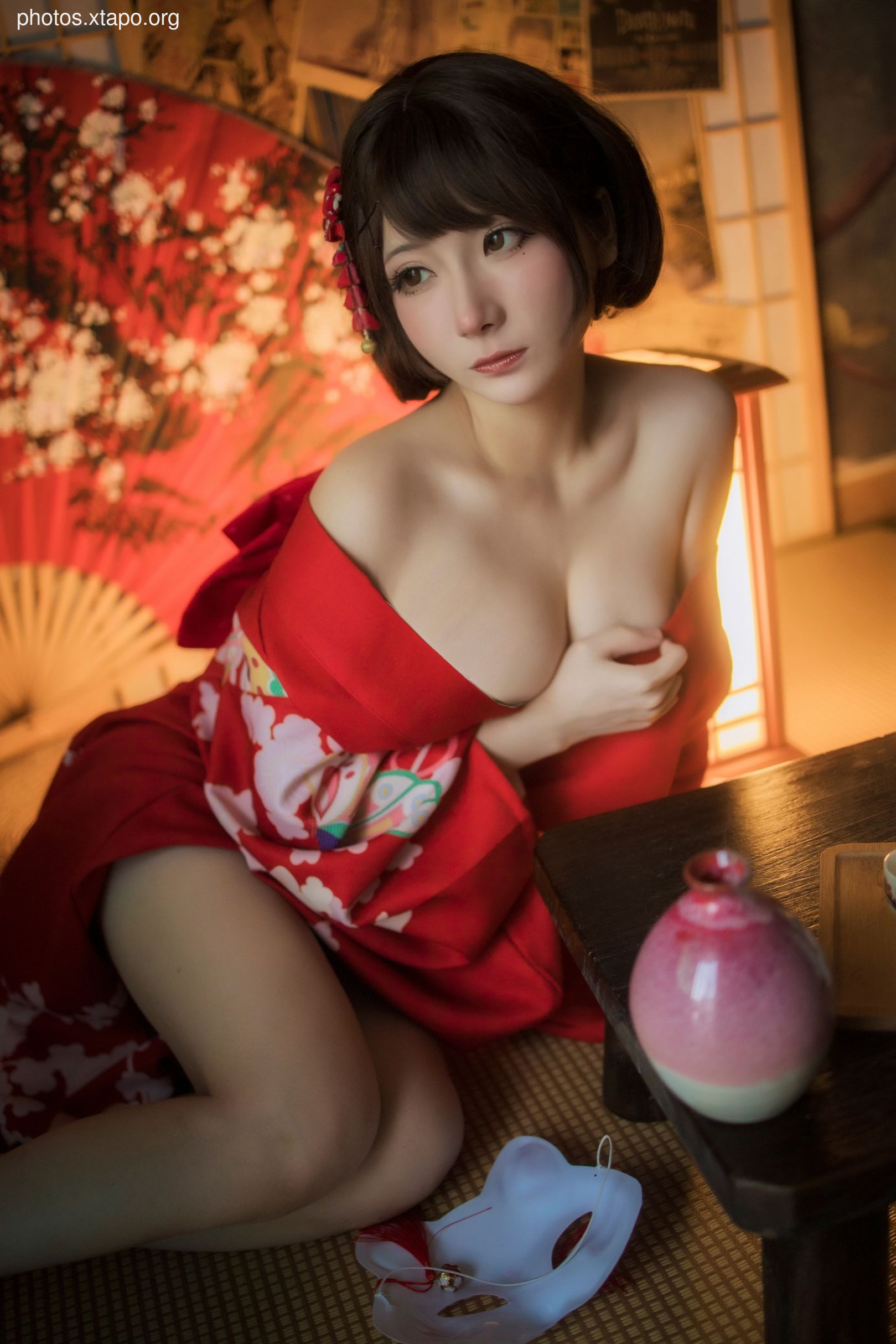 A young woman in a traditional red kimono sits on a tatami mat, holding a mask. The setting is a dimly lit room with Asian decor.