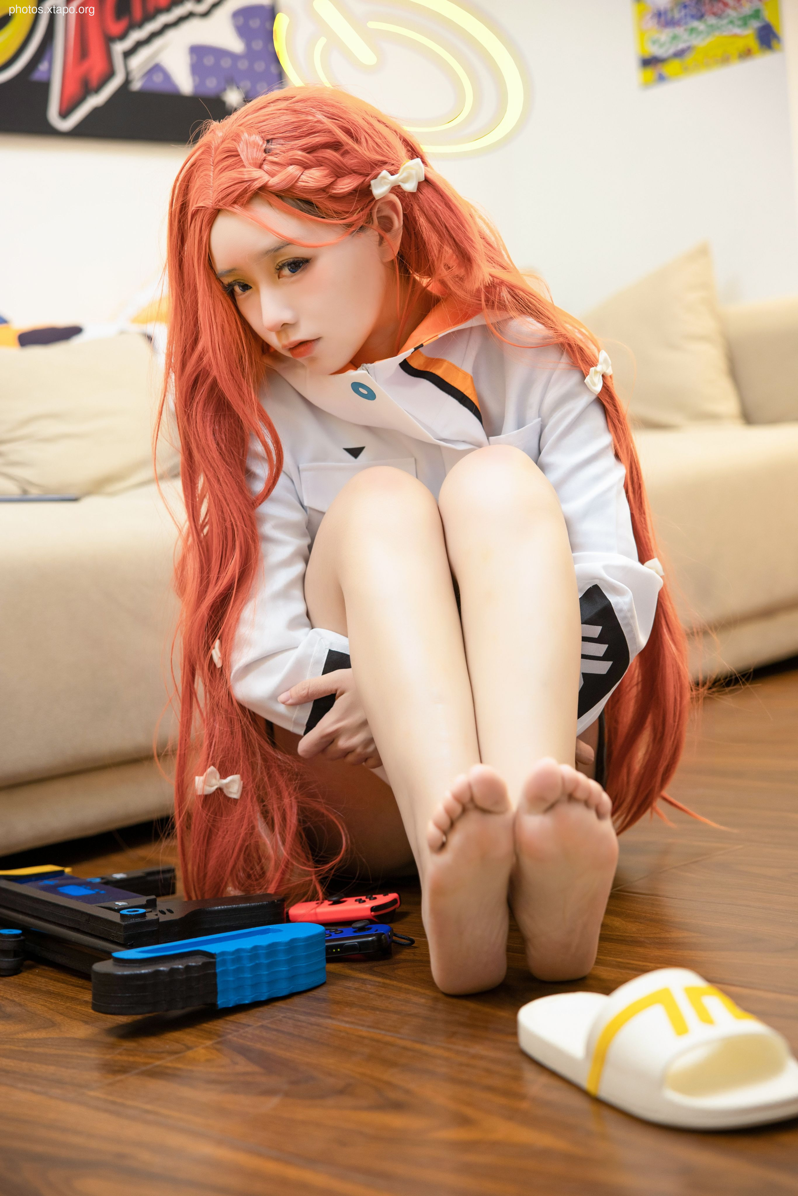 A young woman cosplaying with long orange hair sits on the floor, her legs crossed. She wears a white shirt with orange accents and has white bows in her hair. She is holding a game controller and has a pair of slides nearby.