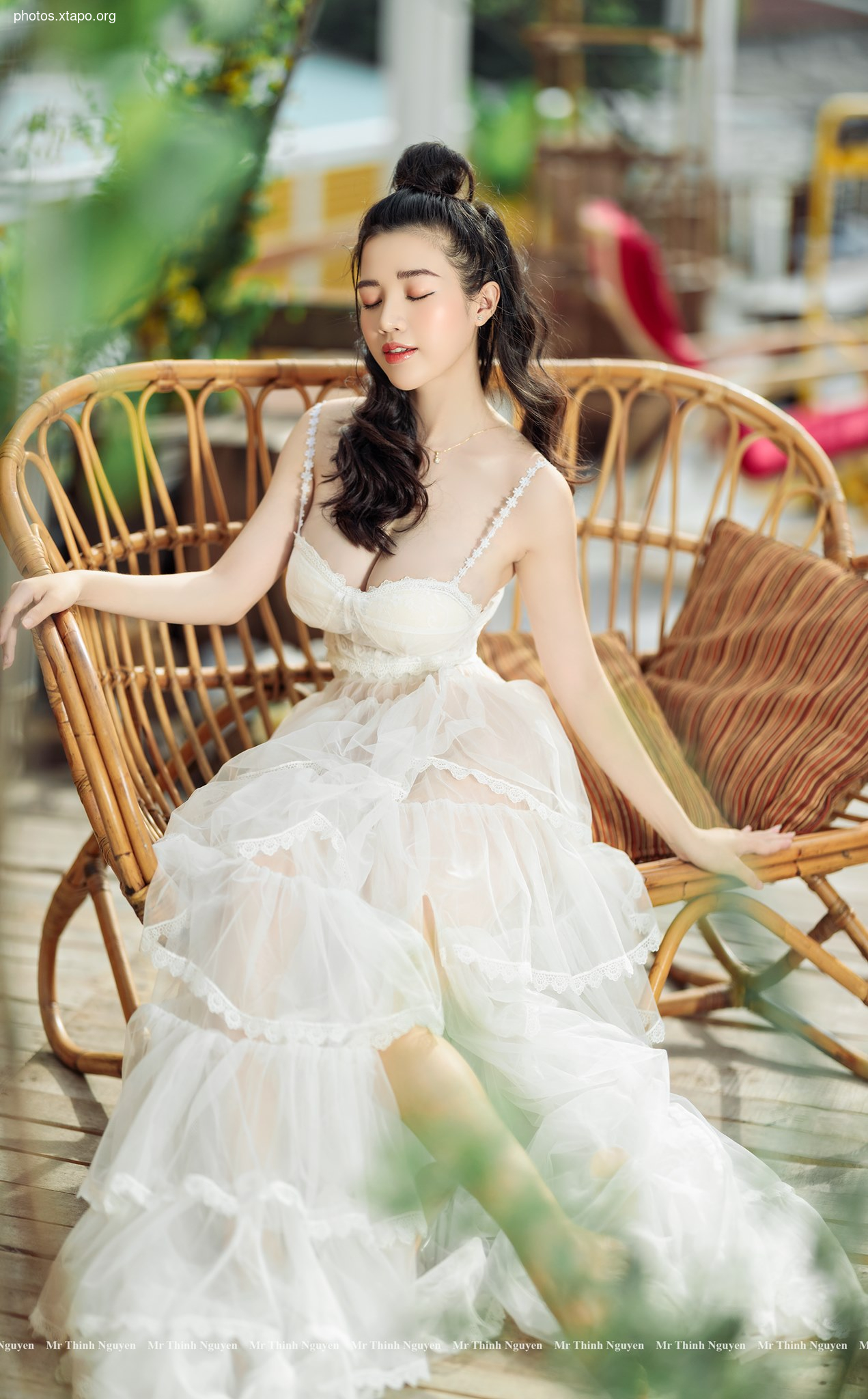 A young woman with her hair in a bun sits on a wicker chair, wearing a flowing white dress. The setting appears to be outdoors, with lush greenery in the background.